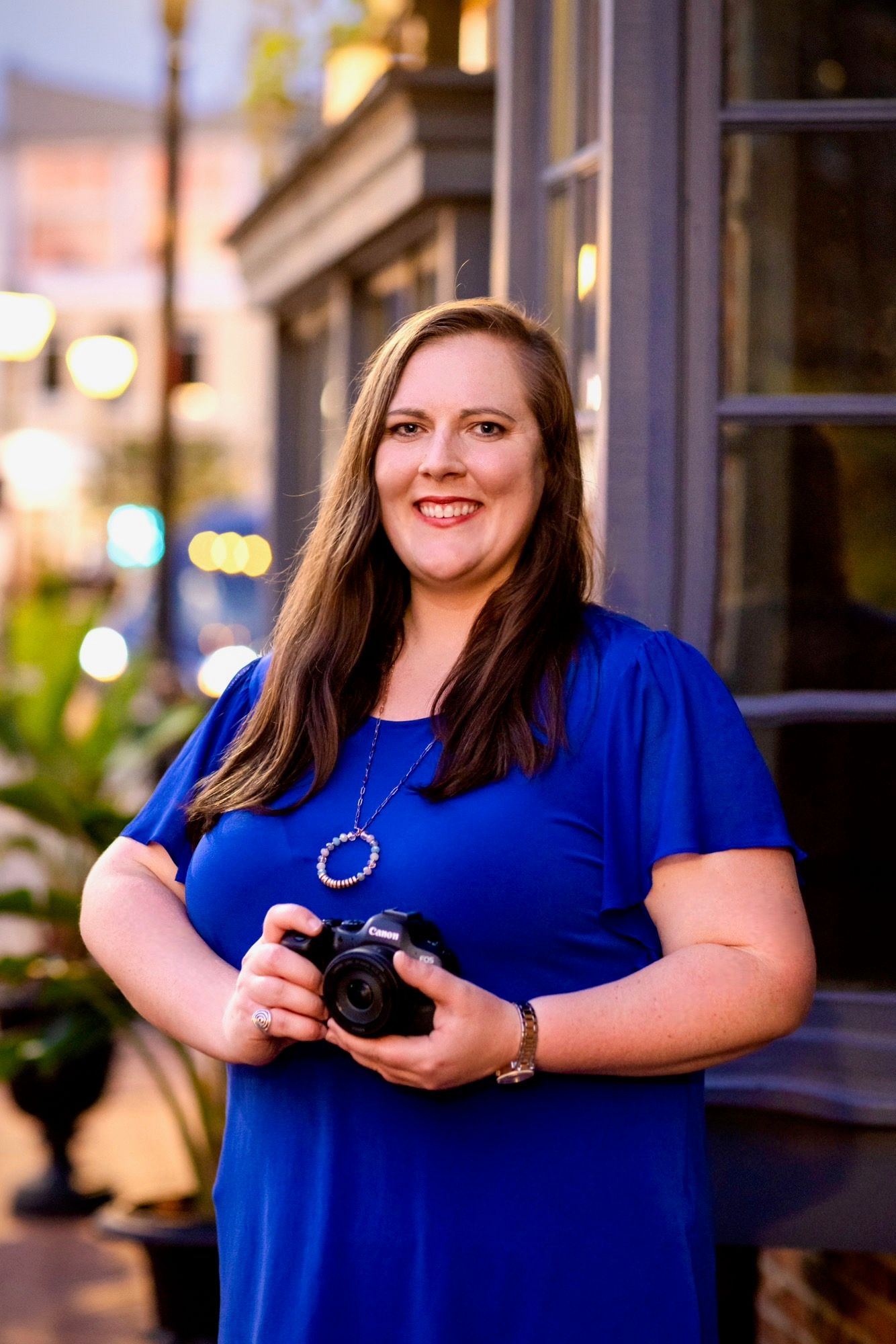 Smiling woman in blue dress holding DSLR camera outdoors at dusk on city street.