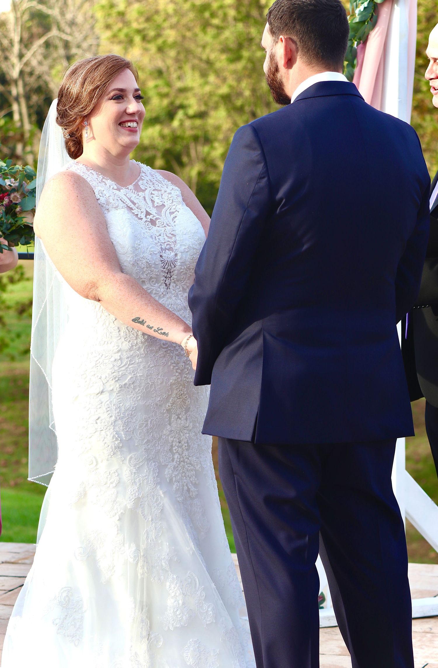 Smiling bride in lace mermaid gown holding hands with groom in navy suit during outdoor wedding ceremony.