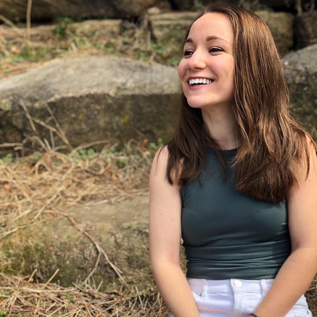 Young woman with brown hair laughing joyfully while sitting on rocks in natural outdoor setting.