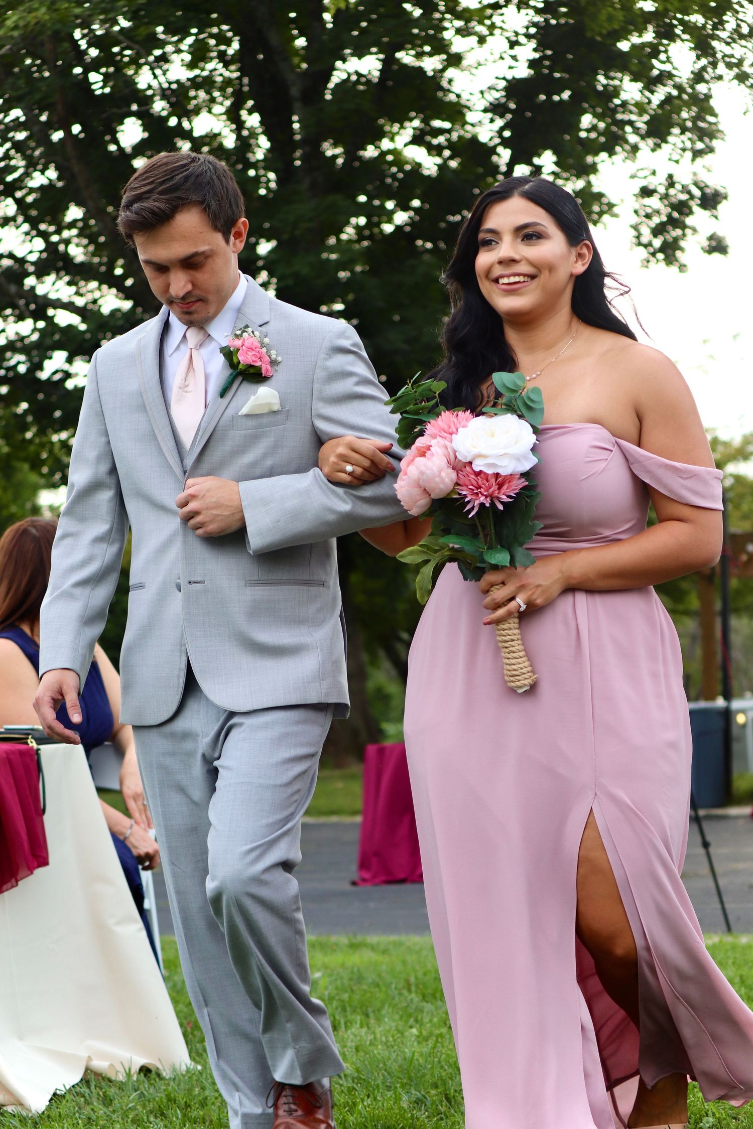 Groomsman in gray suit escorting bridesmaid in dusty rose off-shoulder gown down outdoor wedding ceremony aisle.