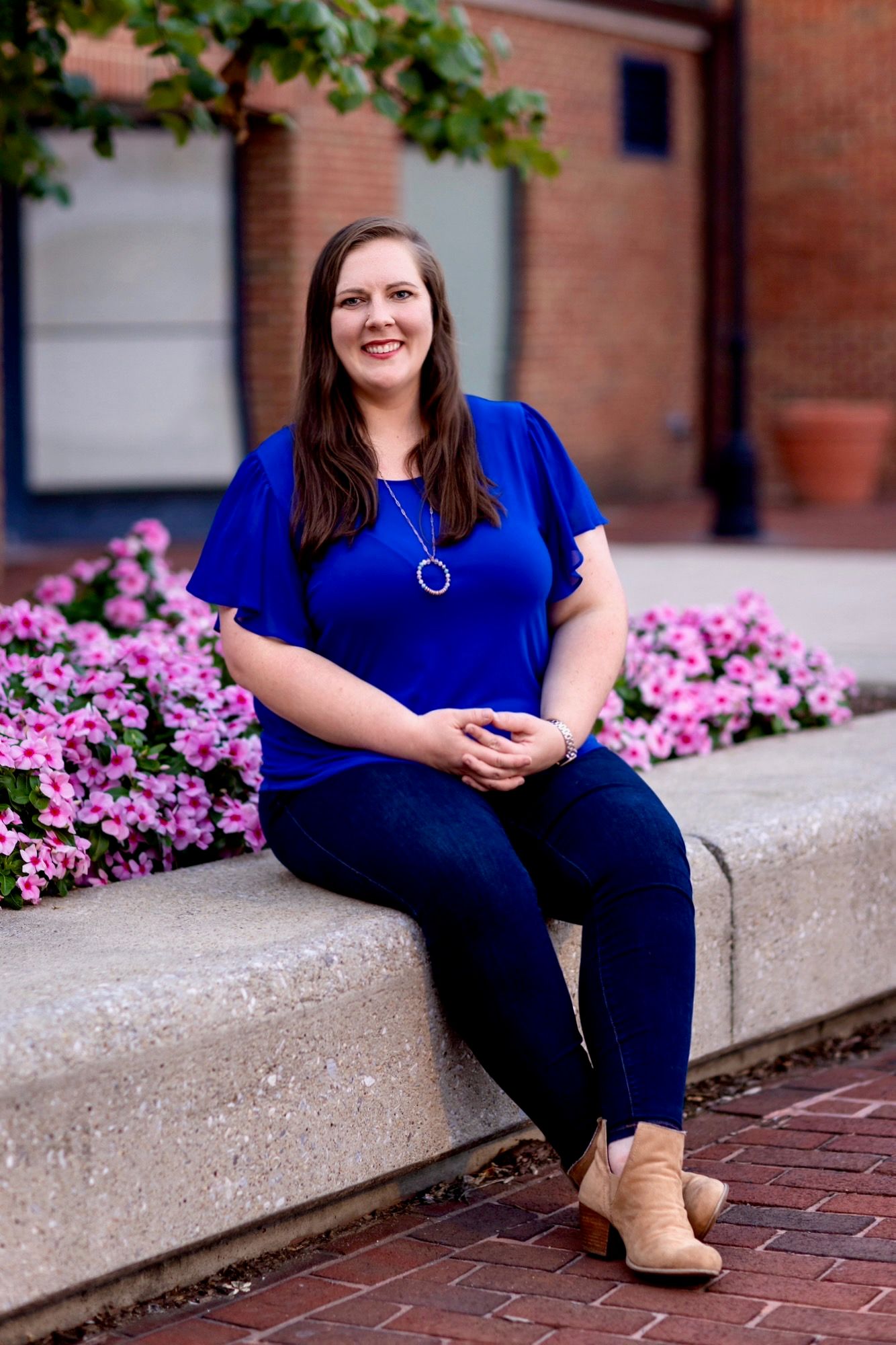 Smiling woman in blue top and jeans sitting on stone ledge with pink flowers.