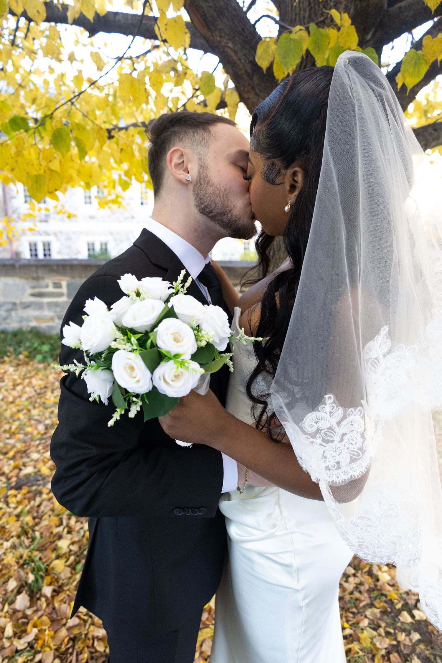 Interracial couple sharing a romantic kiss under autumn yellow foliage on their wedding day.