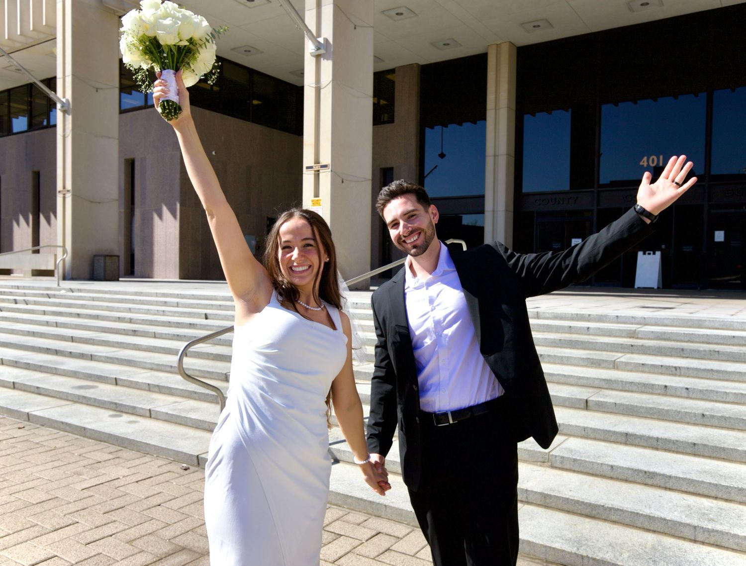Happy newlyweds celebrating outside courthouse steps waving hands holding white bouquet.