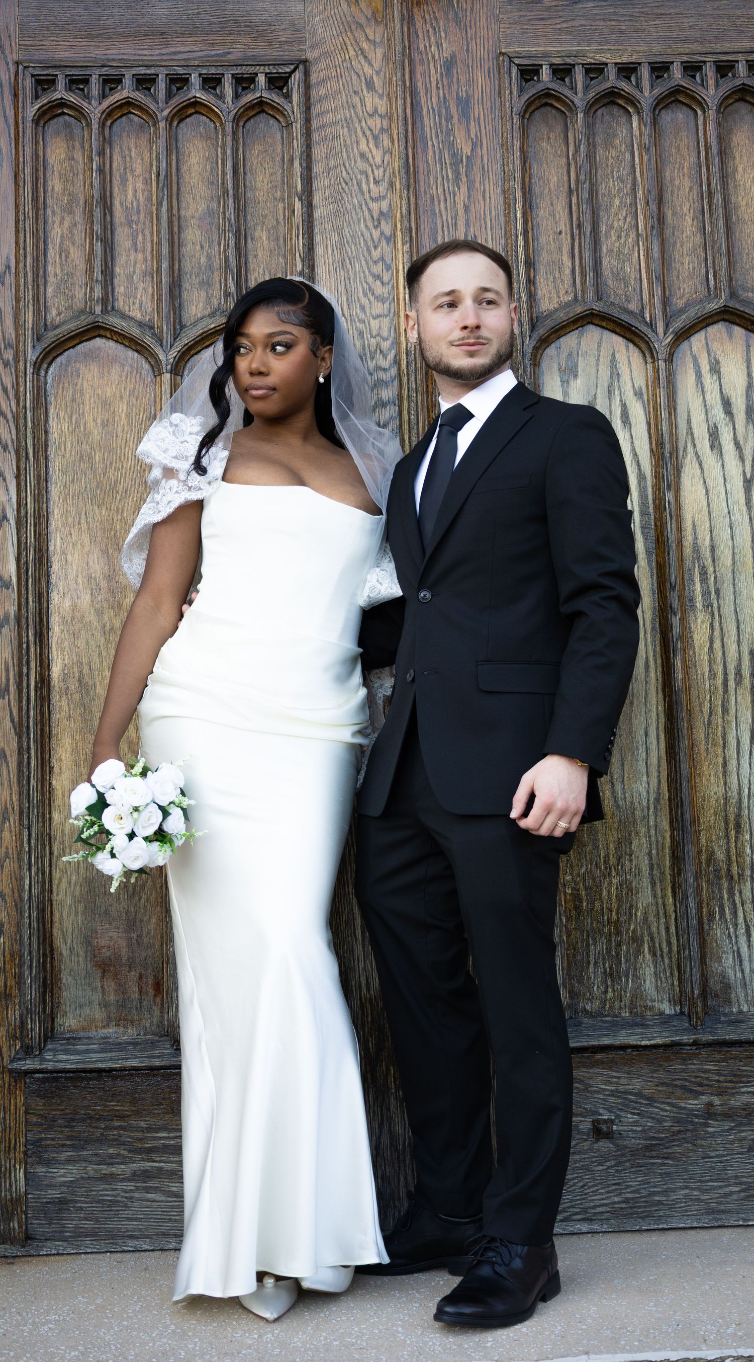 Elegant bride in white off-shoulder gown and groom in black suit posing before ornate doors.