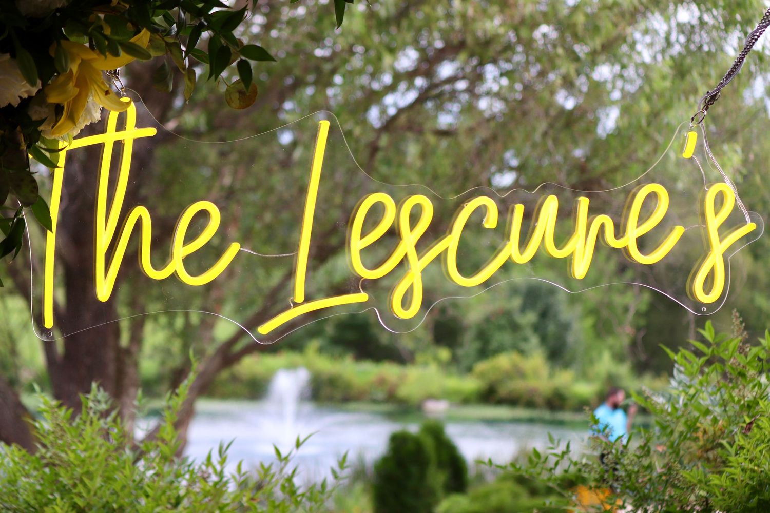 Yellow neon sign reading The Lescures hanging outdoors among green trees near a serene pond.