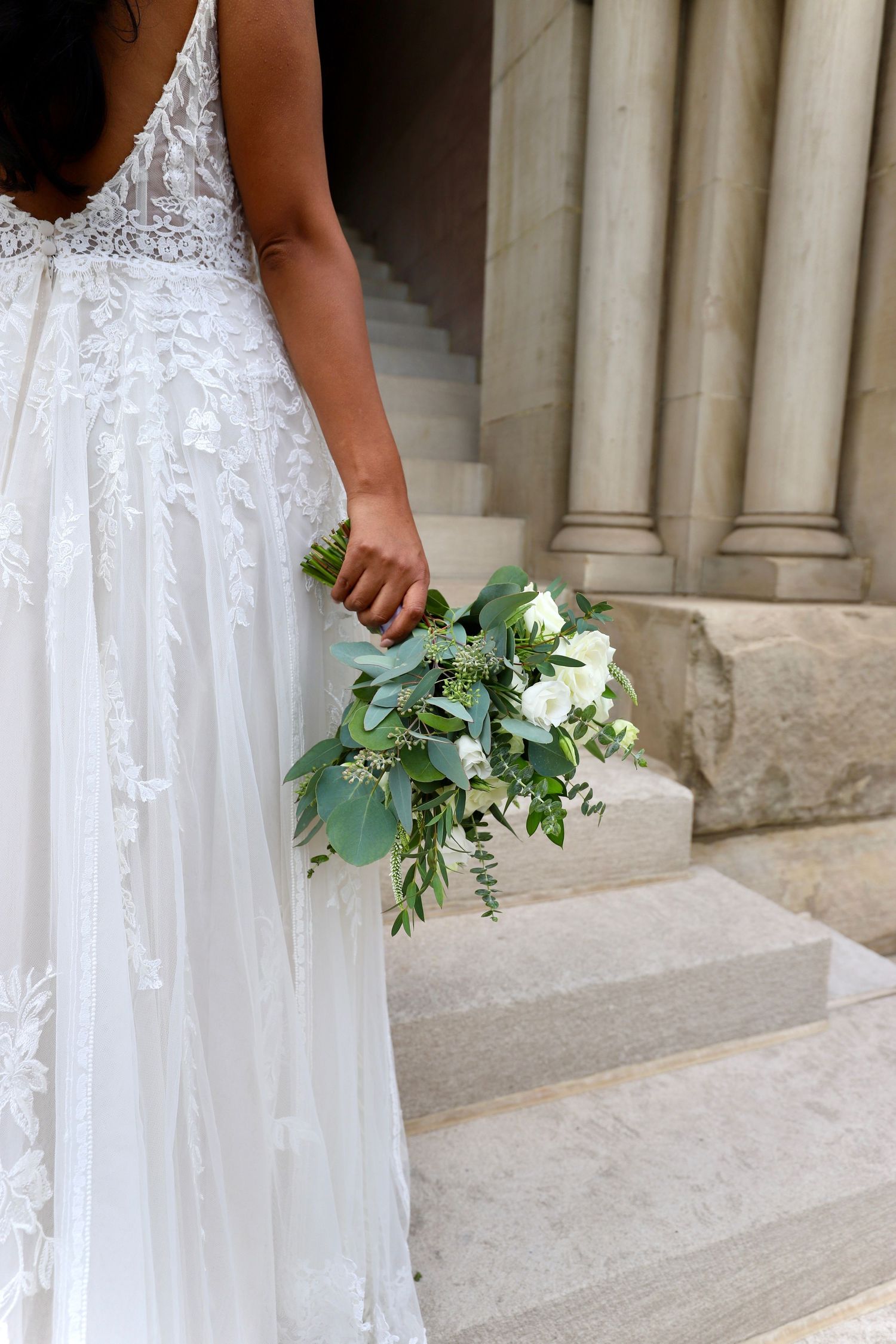 Bride in lace wedding gown holding white and green bouquet on marble steps.