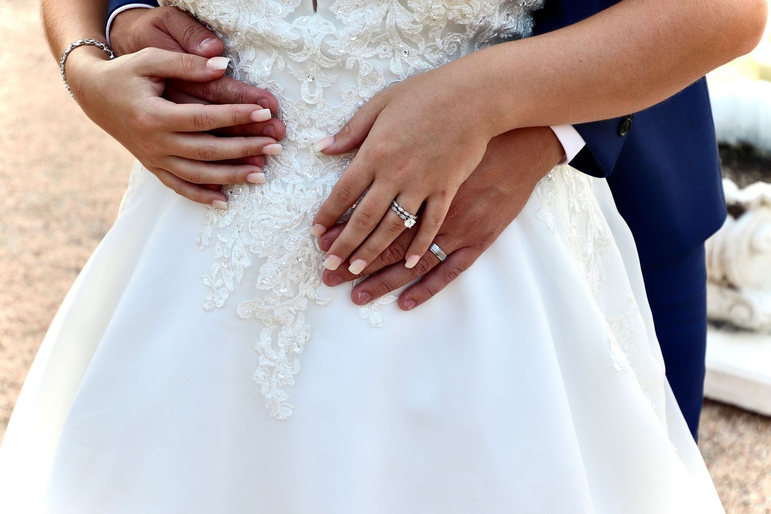 Close-up of bride and groom hands on white lace wedding gown showing diamond engagement and wedding rings.