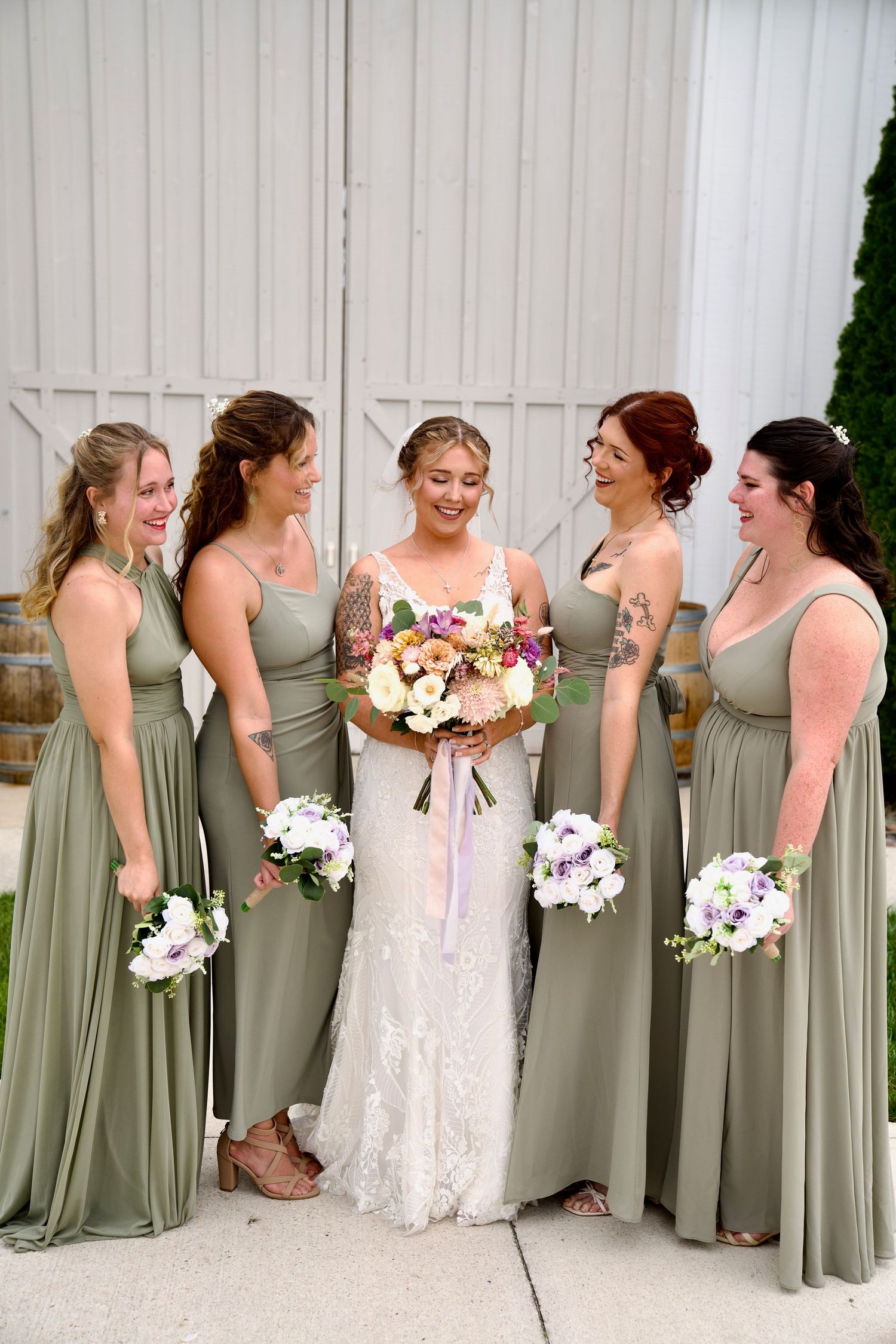 Bride with four bridesmaids in sage green dresses holding bouquets outside a white barn.