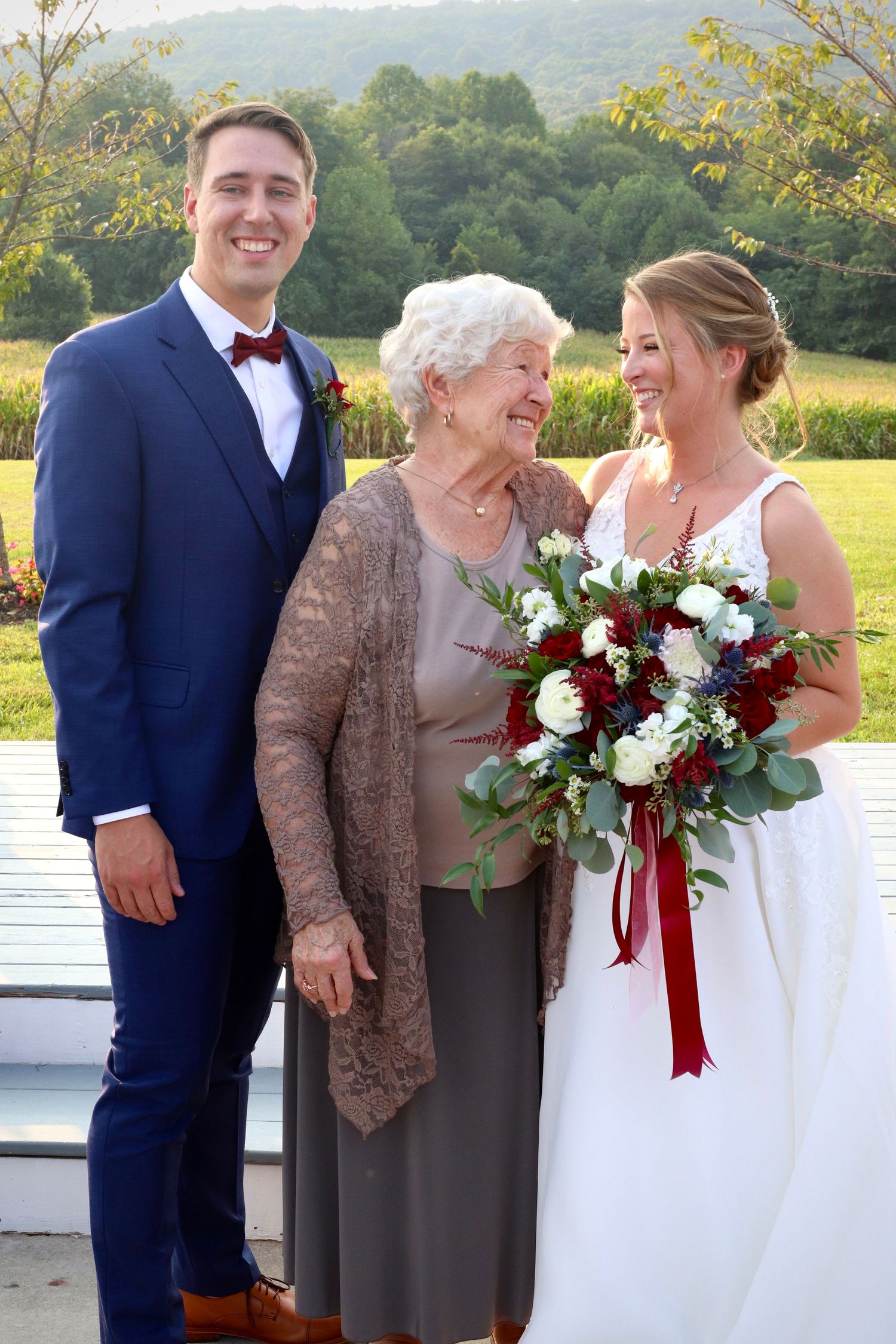 Bride holding red and white bouquet with groom in navy suit posing with elderly woman outdoors.