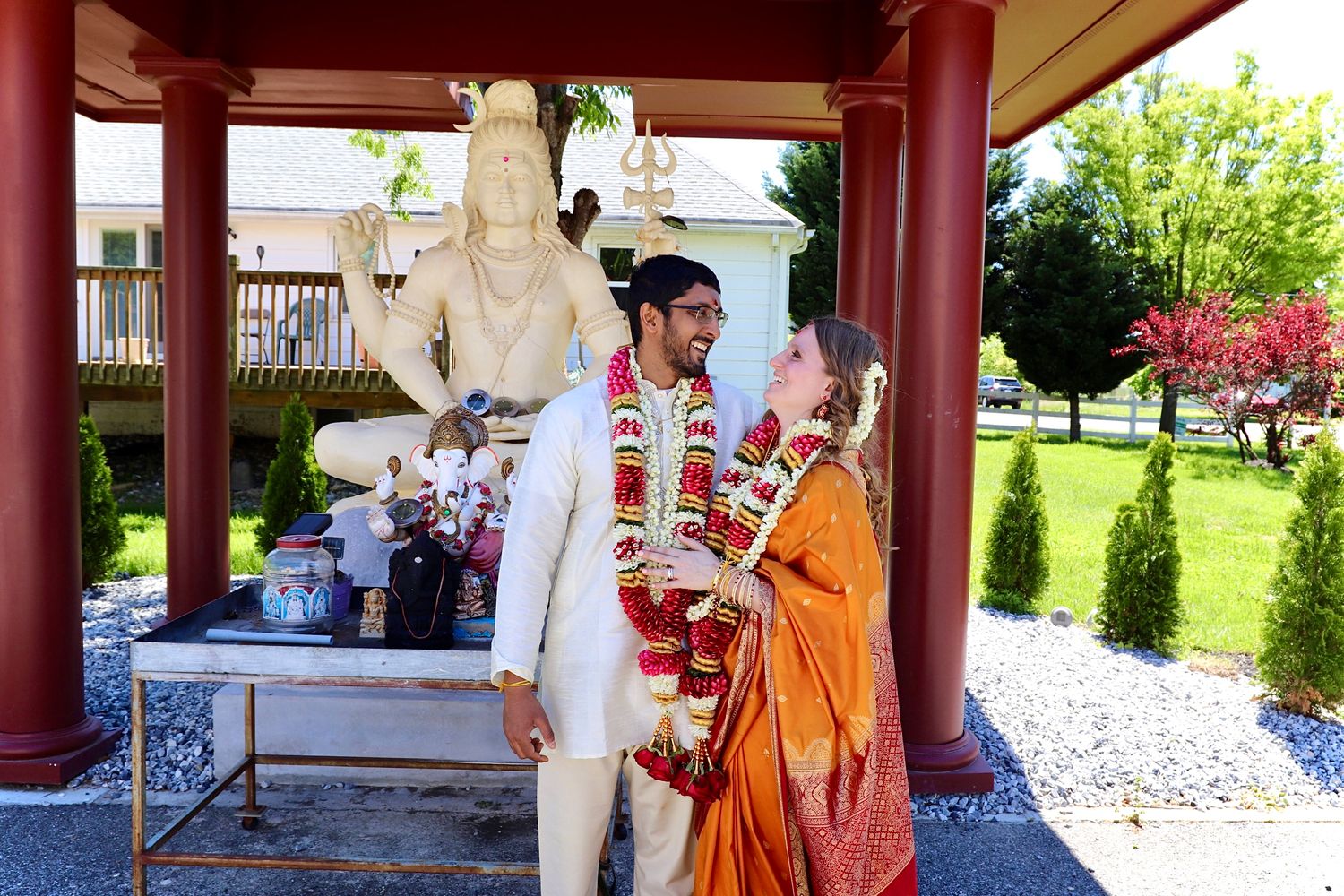 Couple in traditional Indian wedding attire smile together beneath ornate statue during Hindu wedding ceremony outdoors.