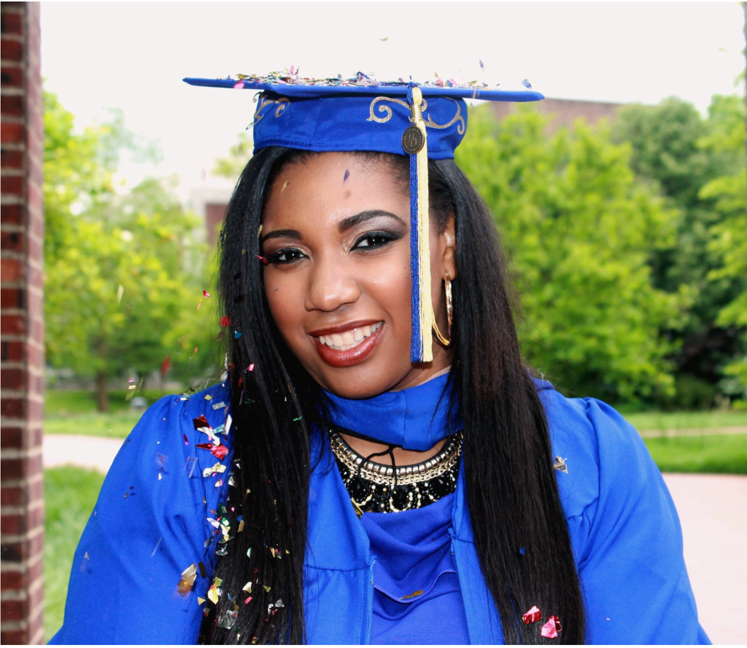 Smiling graduate in royal blue cap and gown with confetti celebrating commencement outdoors on campus.