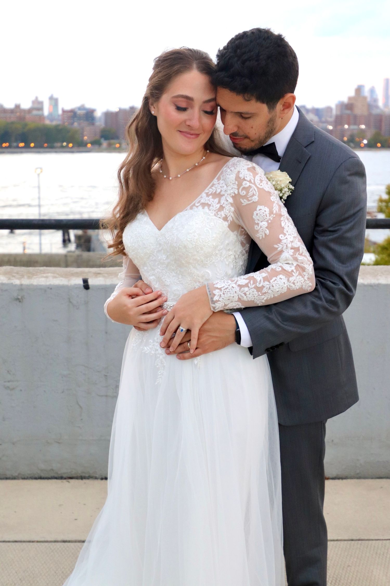 Bride in lace long-sleeve gown embraced by groom in gray suit with city skyline backdrop.