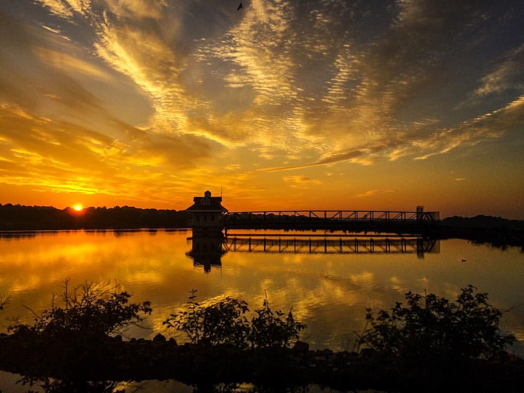 Golden sunset reflecting over calm water with silhouetted pier and building under dramatic cloudy sky.
