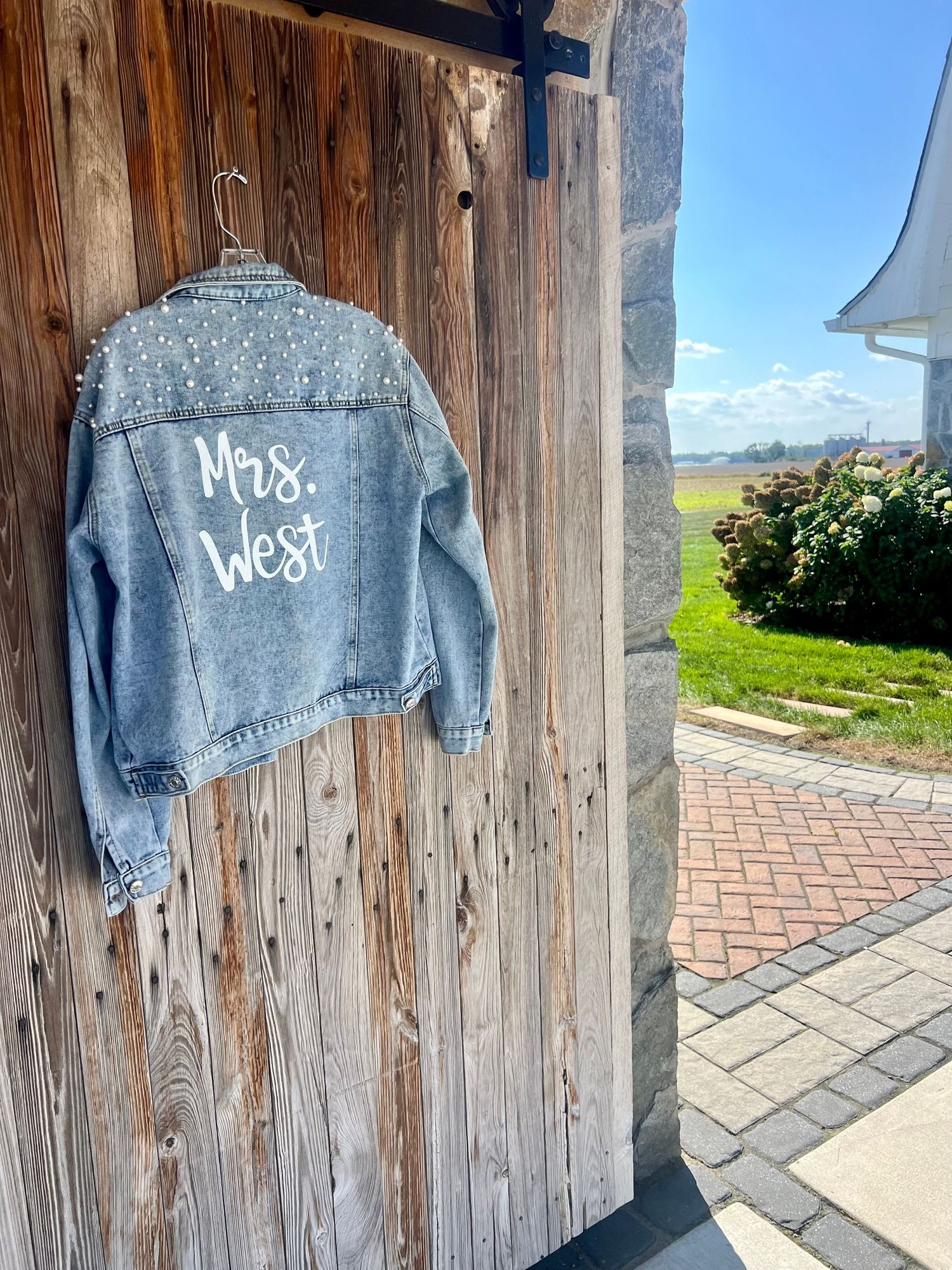 Custom pearl-embellished denim jacket reading Mrs. West hanging on rustic barn door at outdoor wedding venue.