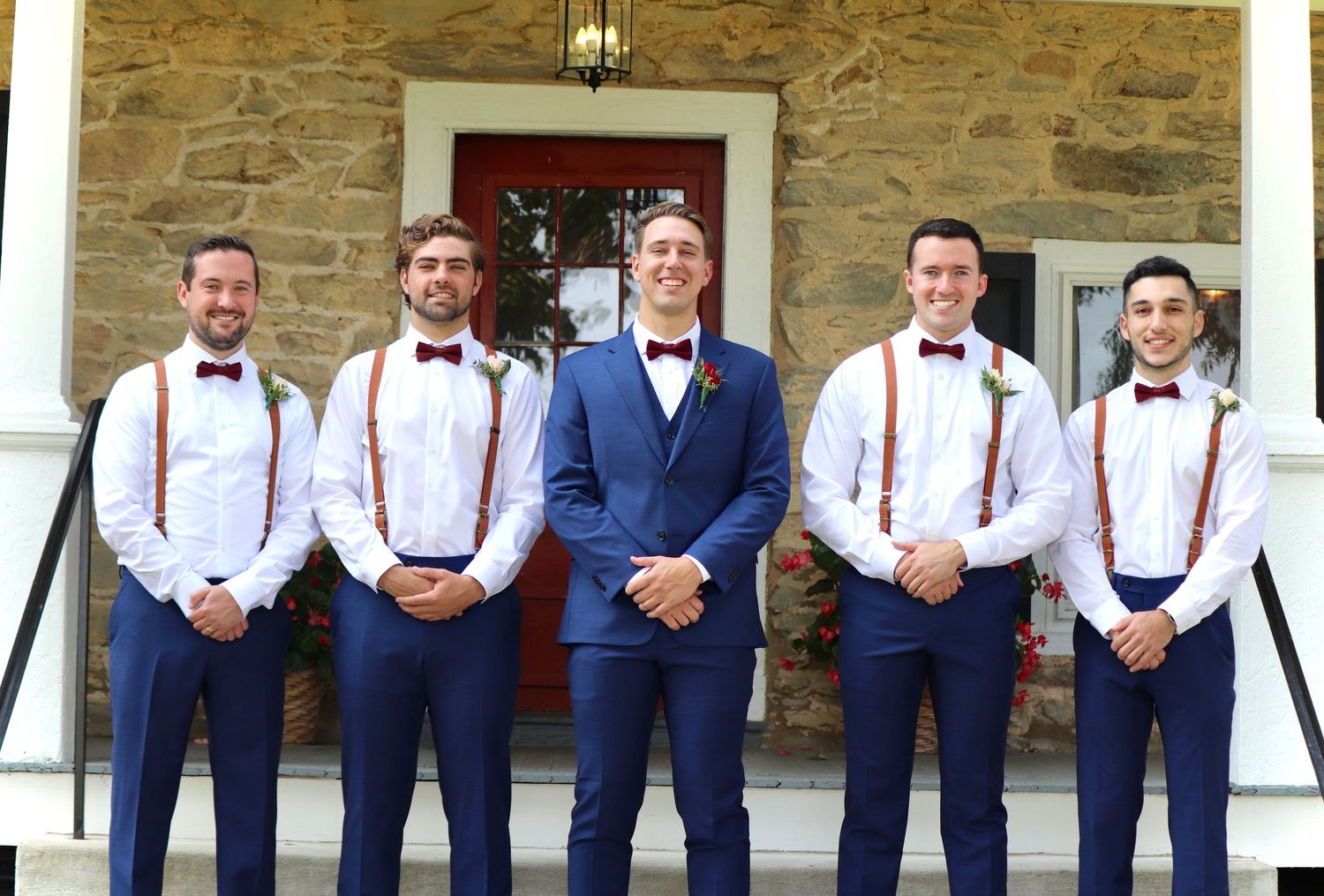 Groom in blue suit with groomsmen wearing white shirts, suspenders, and bow ties posing on stone building steps.