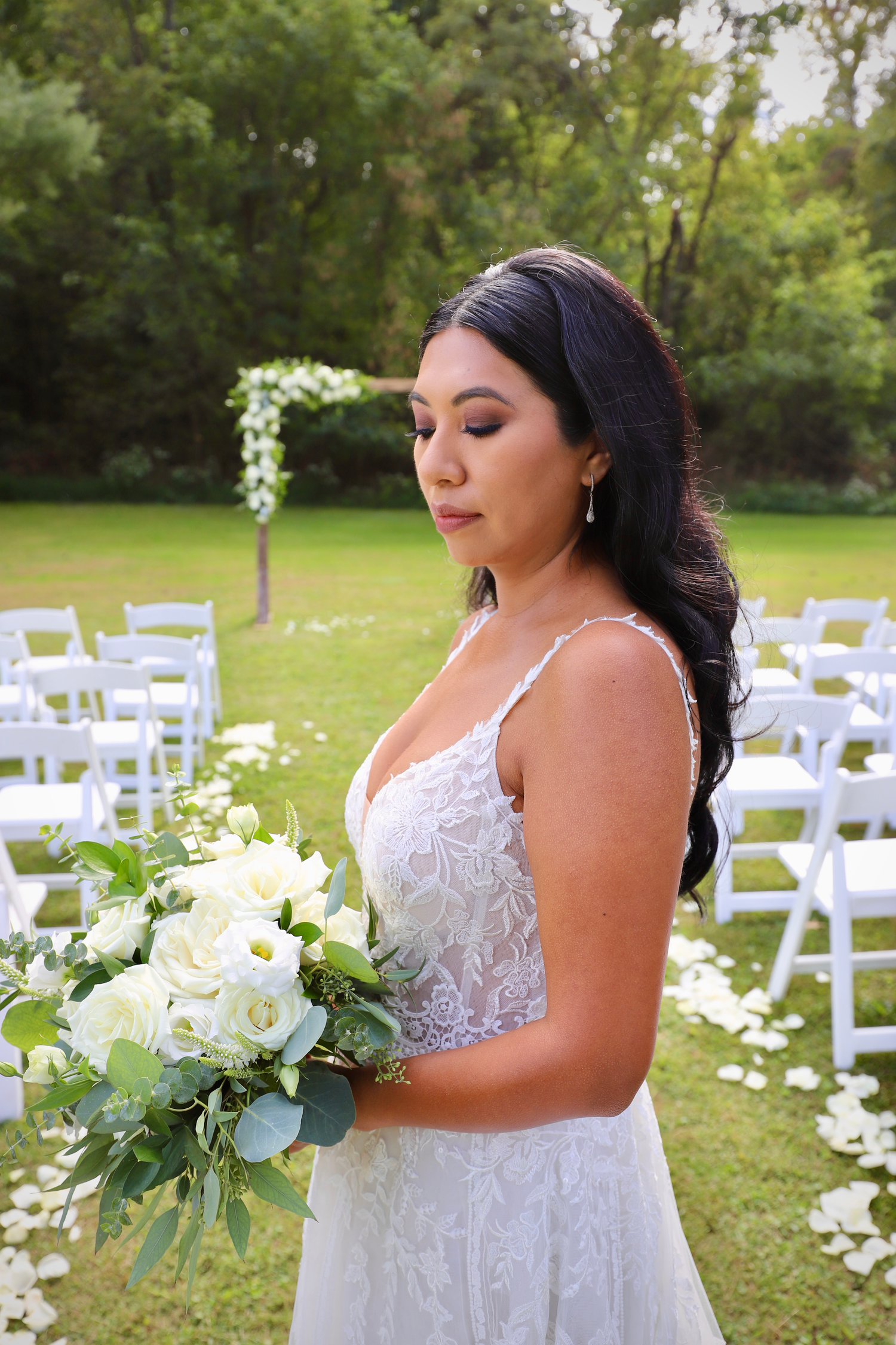 Stunning bride in lace wedding dress holds white bouquet at outdoor garden ceremony venue.