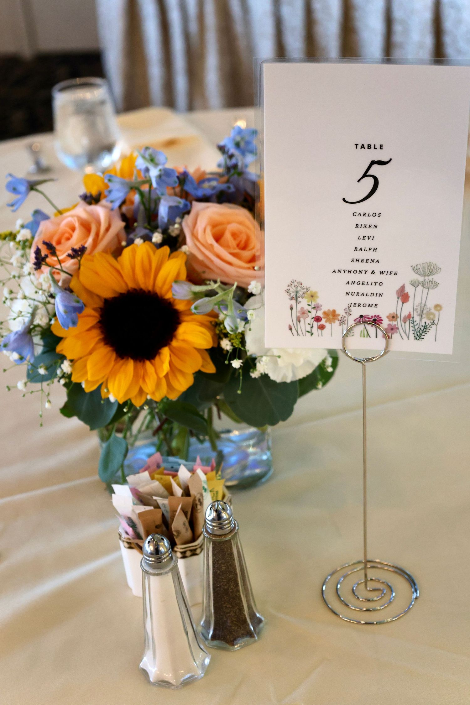 Wedding table centerpiece with sunflowers, pink roses, and blue flowers beside a Table 5 seating card.