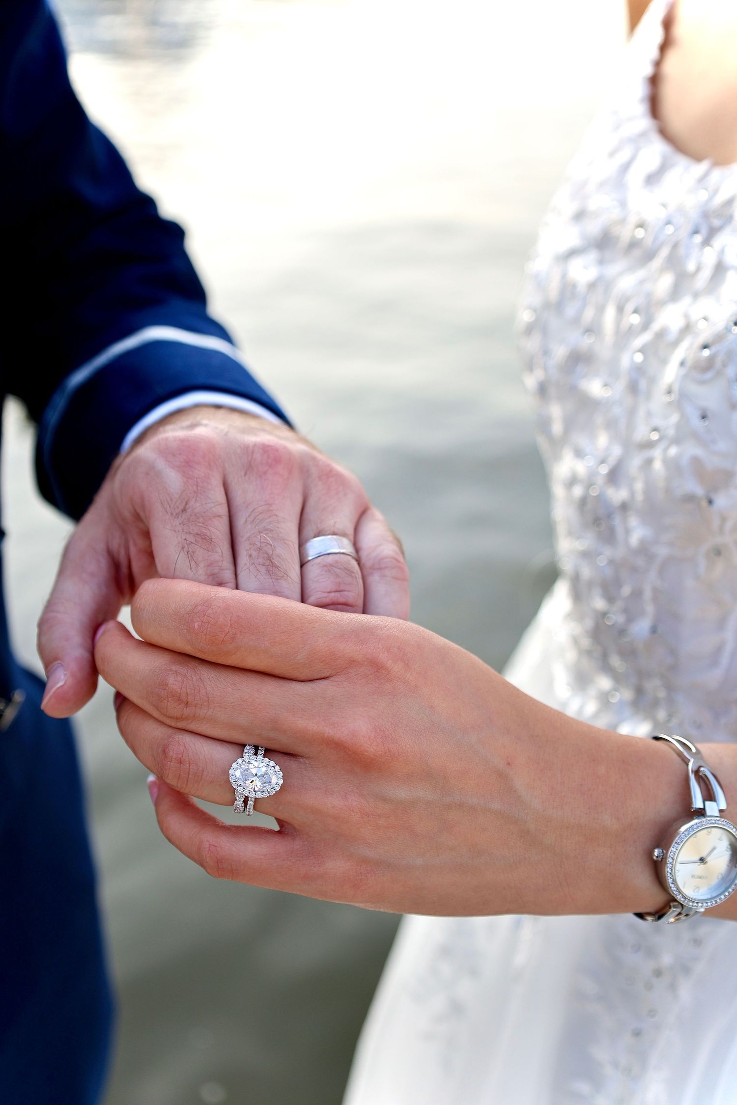 Newlyweds hold hands displaying wedding rings, bride wearing elegant diamond halo ring and pearl bracelet.