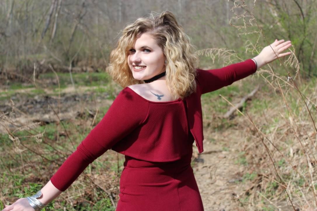 Woman in red dress with curly hair posing outdoors in early spring woodland setting.