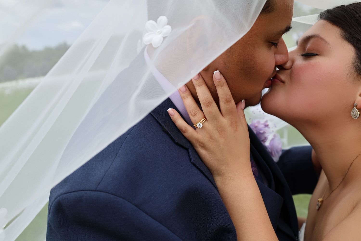 A couple shares a romantic kiss under a wedding veil, showcasing a beautiful diamond engagement ring.