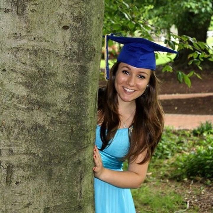 Smiling young woman in blue graduation cap peeking out from behind a tree.