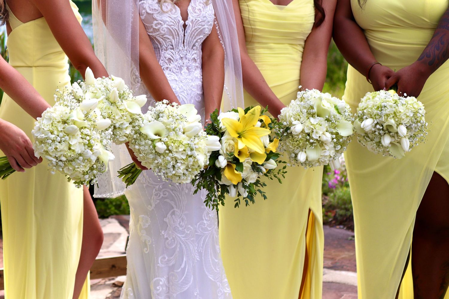 Bride in lace gown with bridesmaids in yellow dresses holding white hydrangea and yellow floral bouquets outdoors.