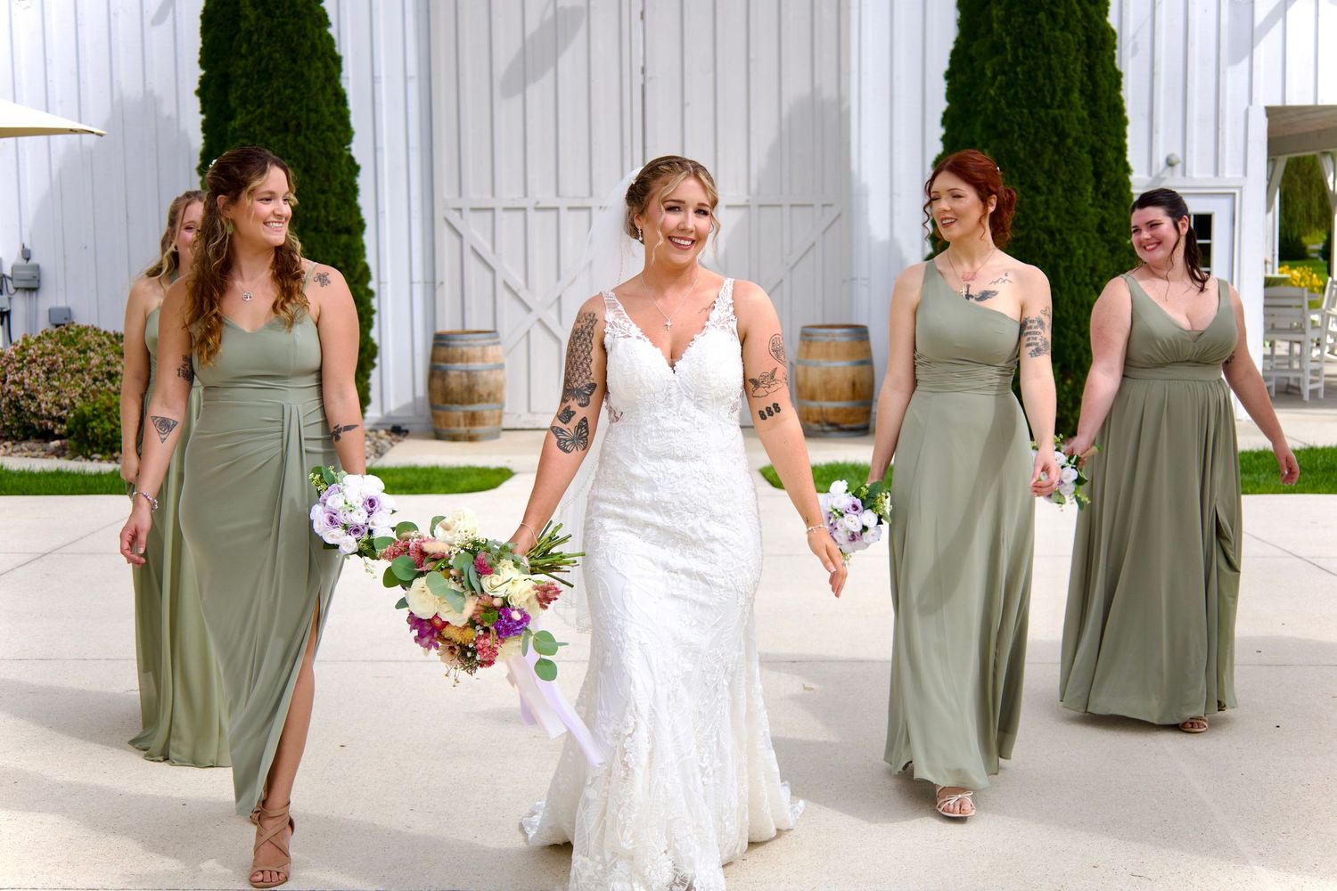 Bride in lace wedding gown walks confidently with three bridesmaids in sage green dresses near white barn venue.