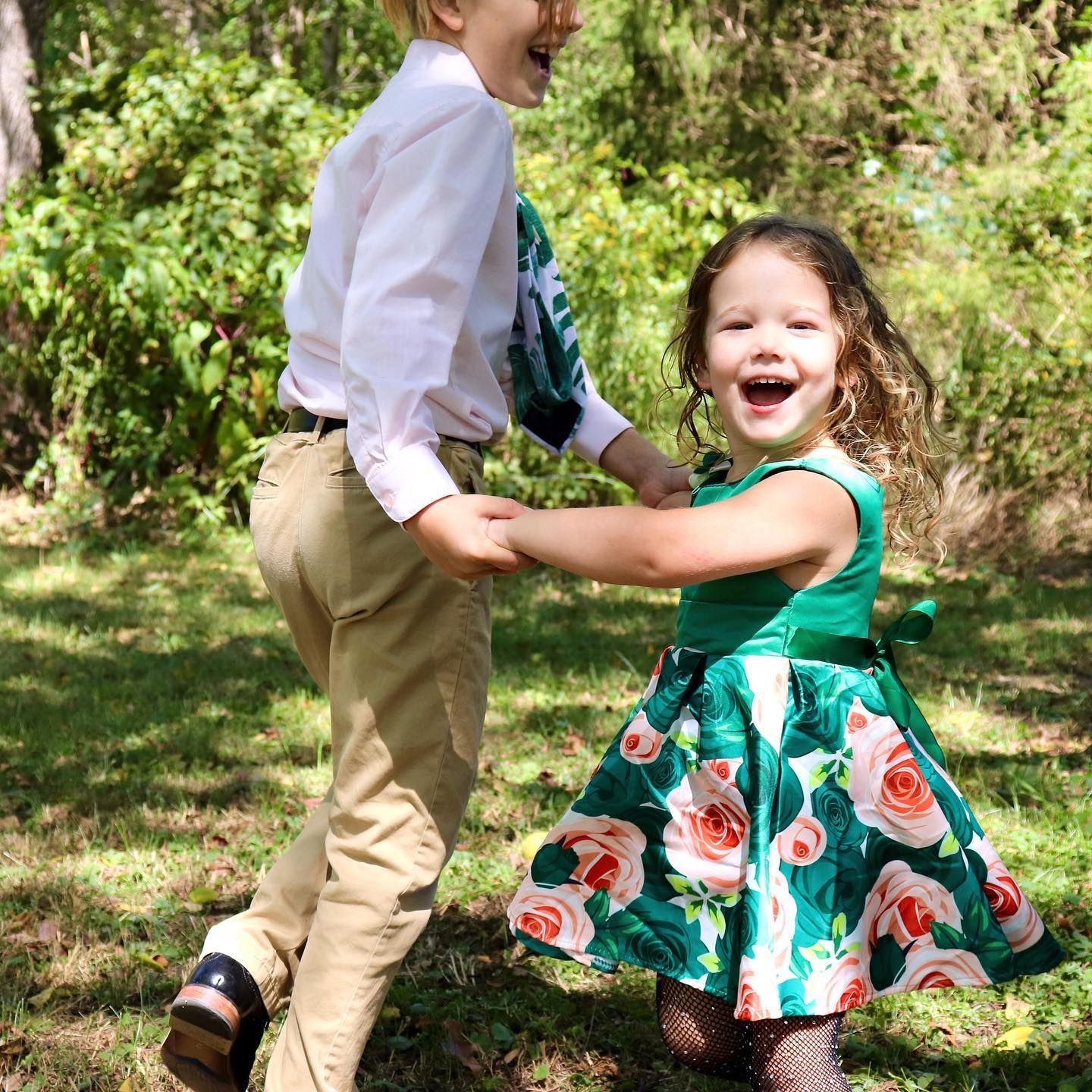Two young children in formal attire laughing and dancing together in green park.