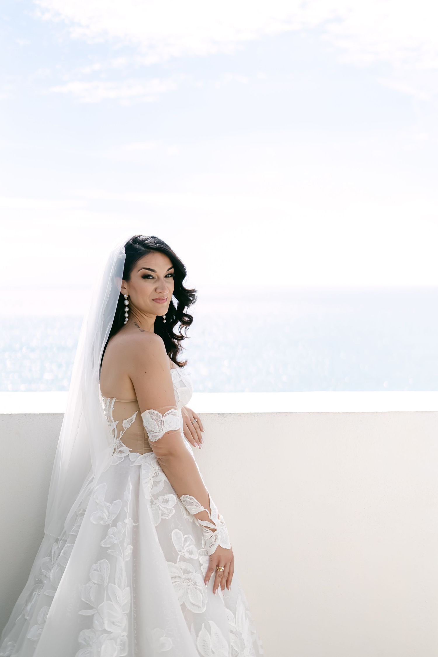 A bride in an elegant off-shoulder lace wedding gown and long veil poses against a bright white background.