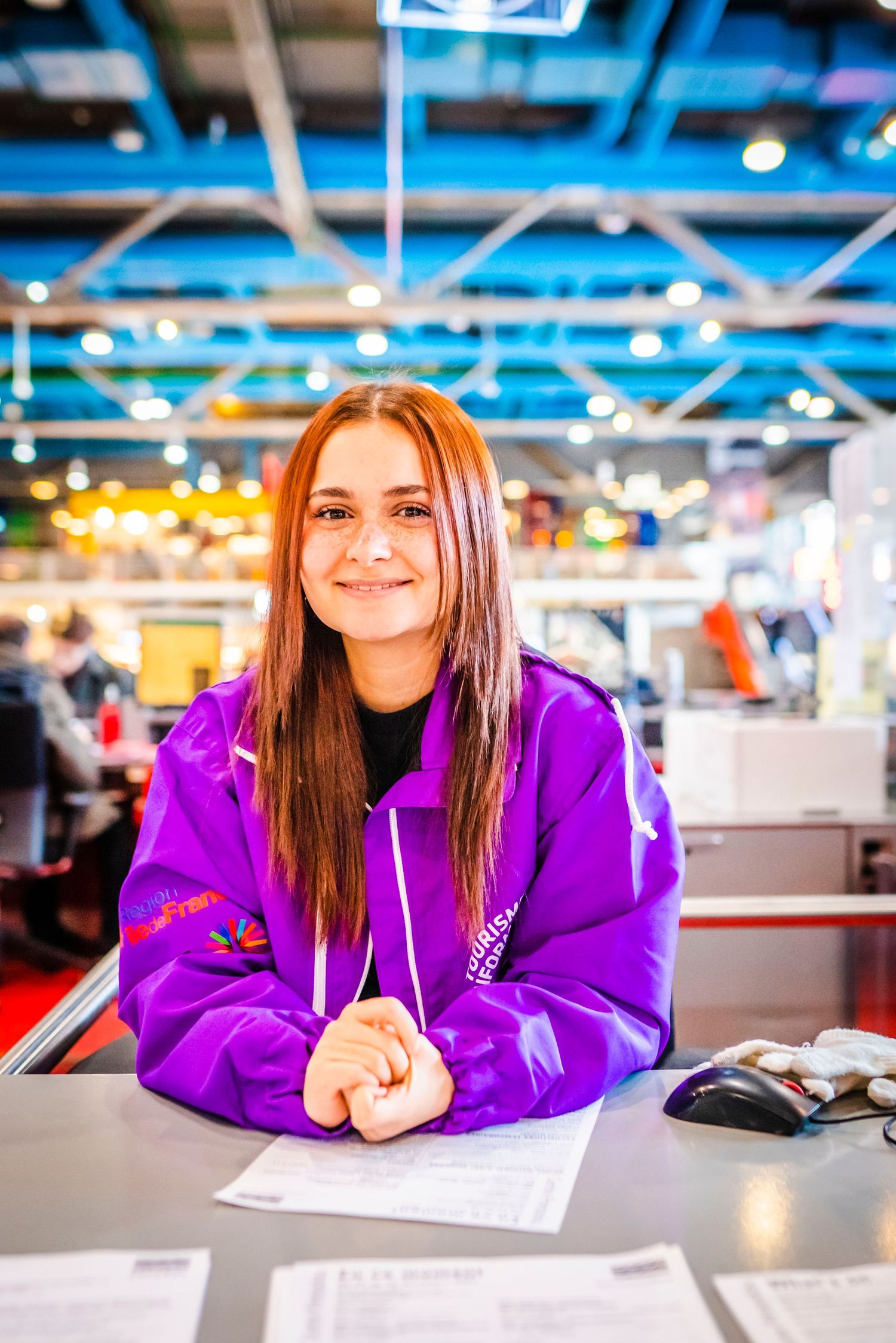 Student wearing bright purple hoodie smiles while sitting at desk with papers in modern indoor setting.