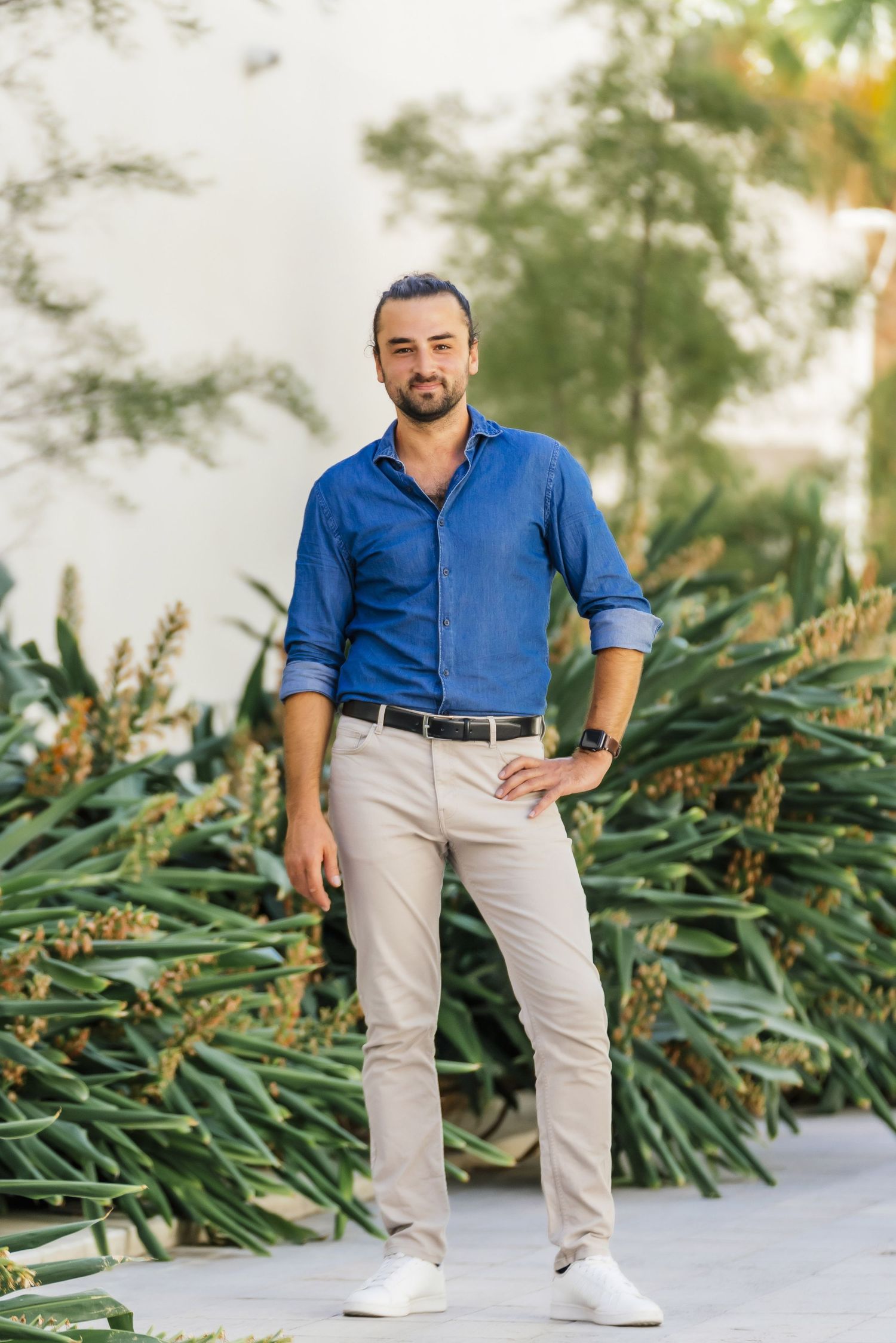 Person wearing a blue button-down shirt and khaki pants standing in a garden setting with tropical plants.