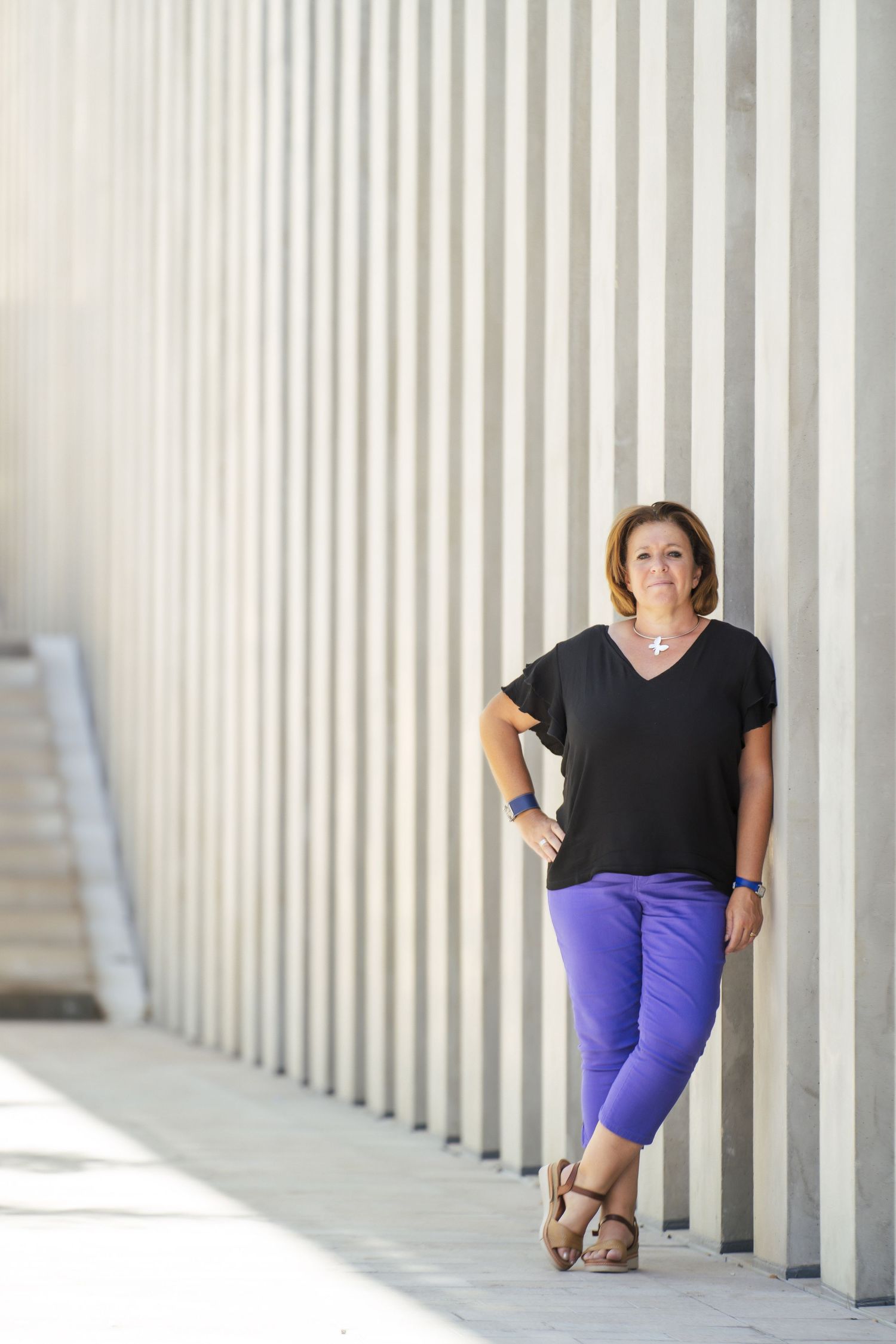 A person in a black v-neck shirt and purple pants poses casually against white architectural columns in natural light.
