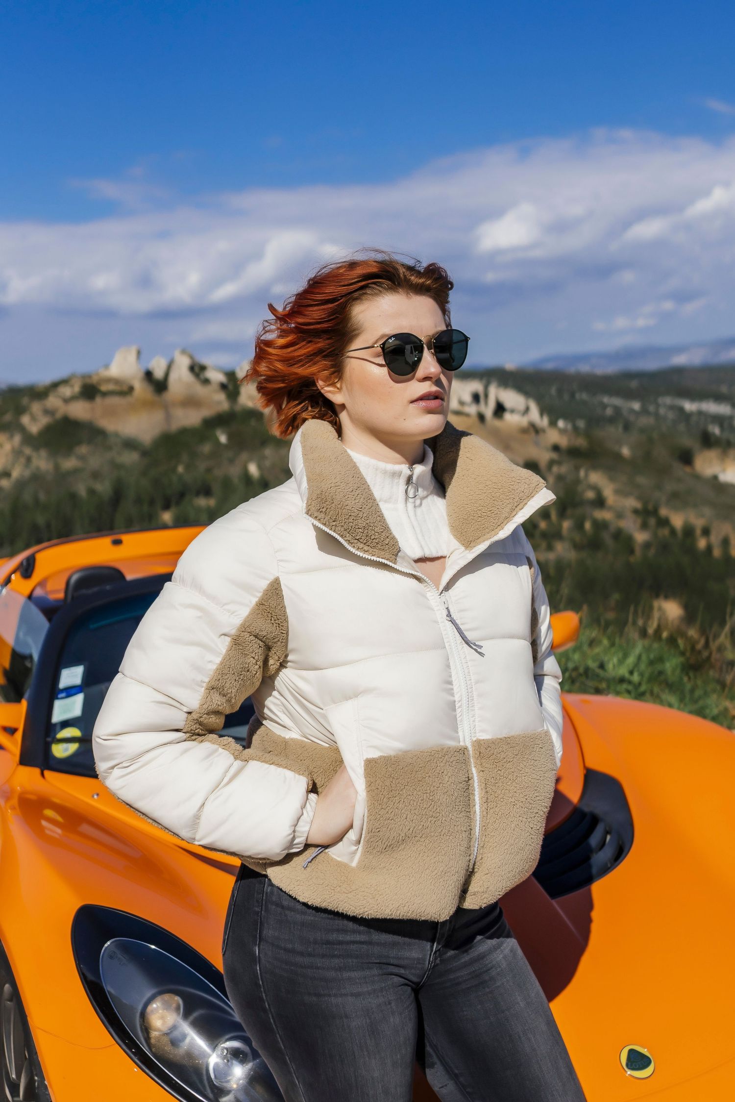 Person in white puffer jacket and sunglasses poses against bright orange sports car with mountain landscape in background.