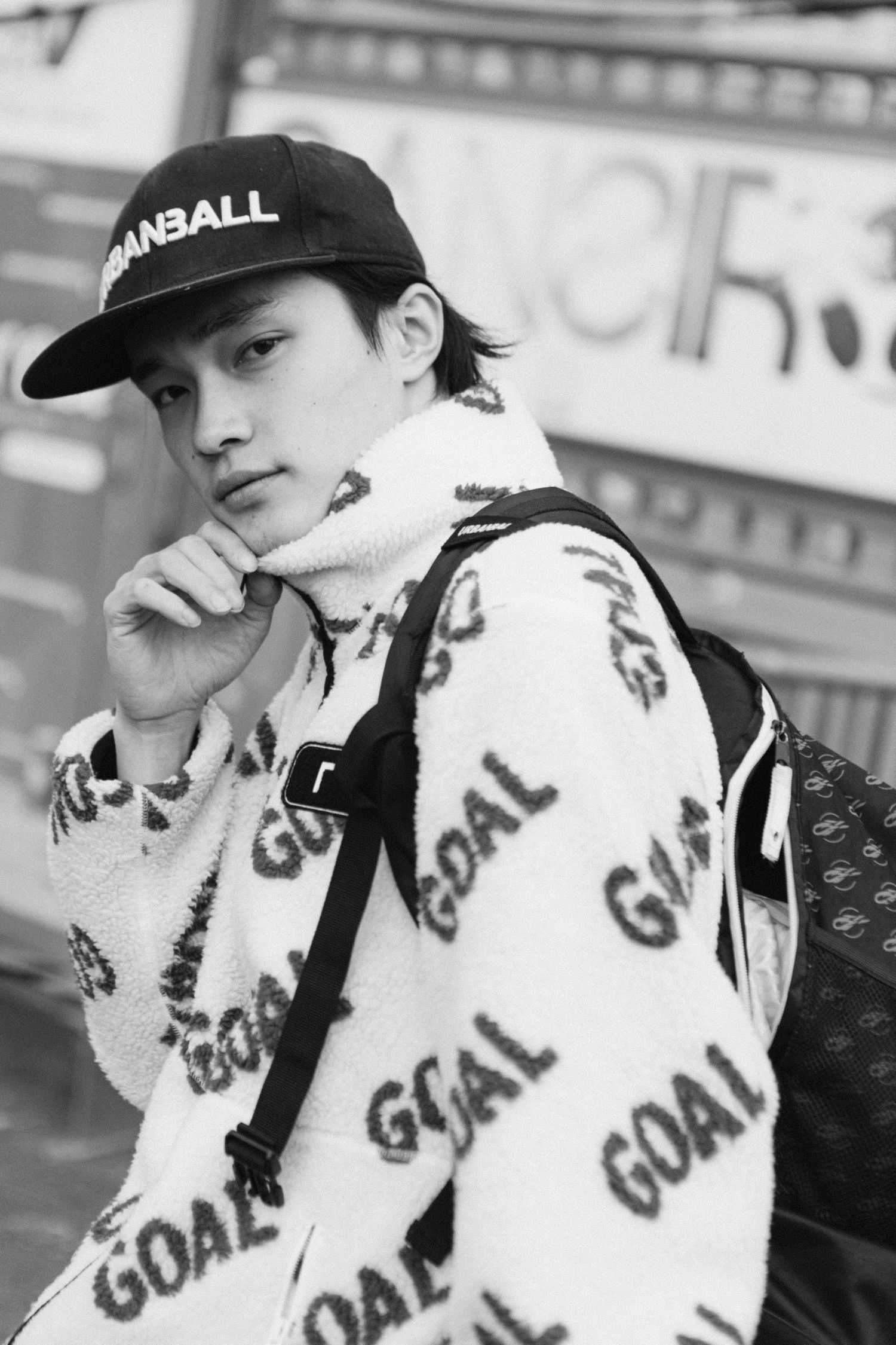 Person wearing a white jacket with 'GOAL' text pattern and black baseball cap in urban setting.