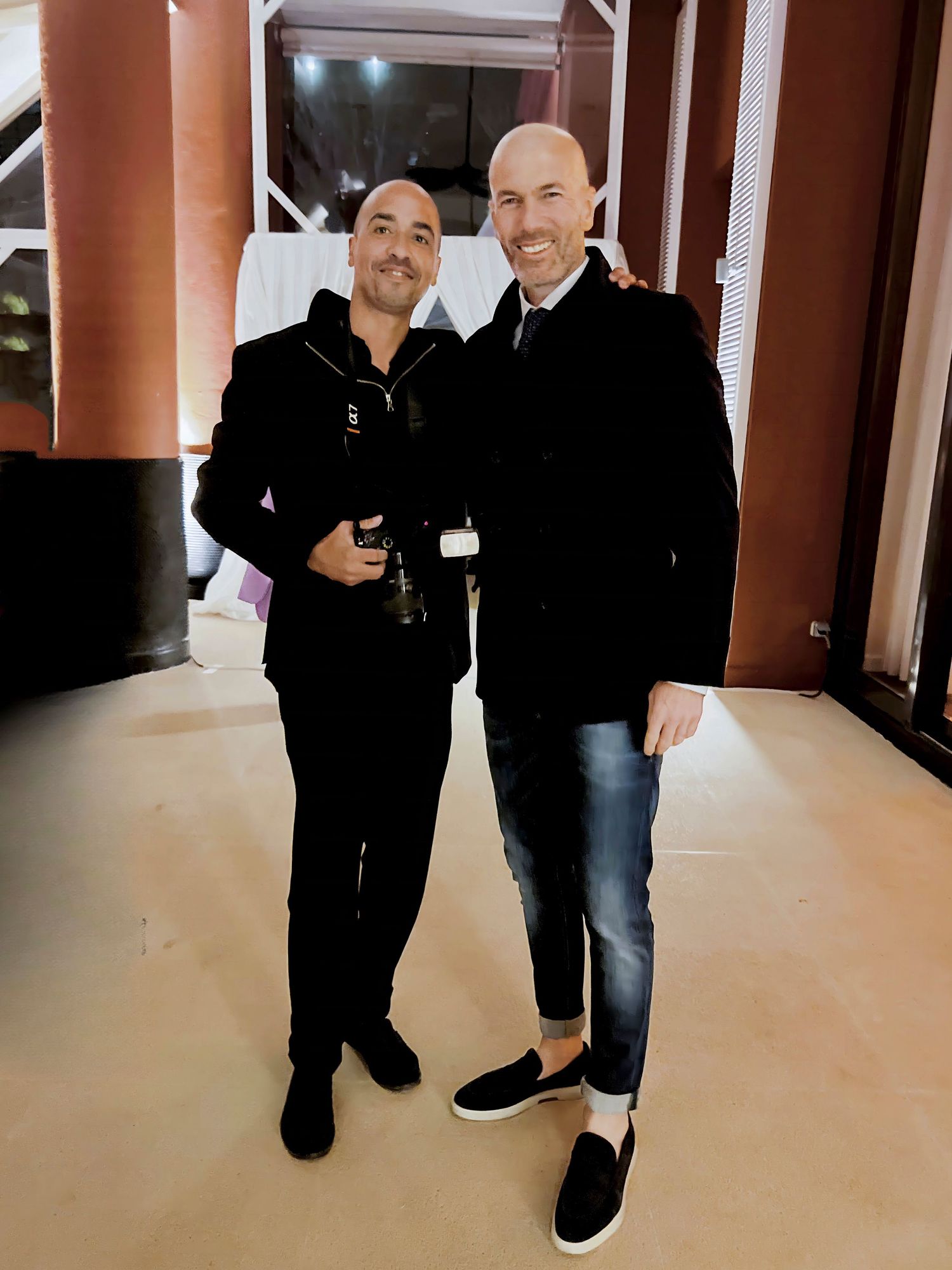 Two men pose together indoors wearing casual attire, one in all black and one in a black coat with jeans.