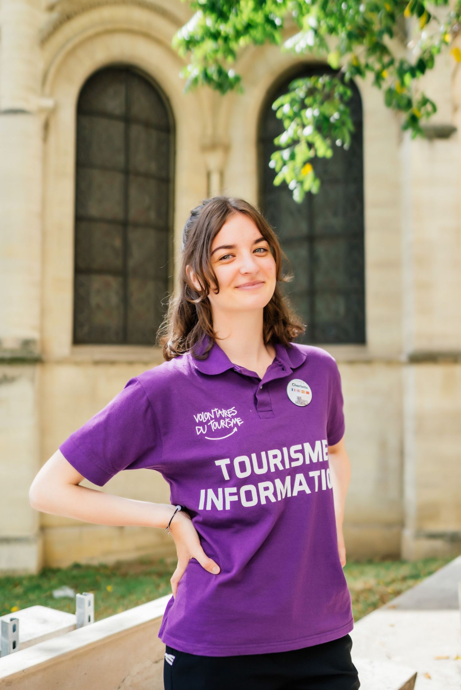 Tourism information staff member wearing a purple polo shirt stands in front of an arched stone building with windows.