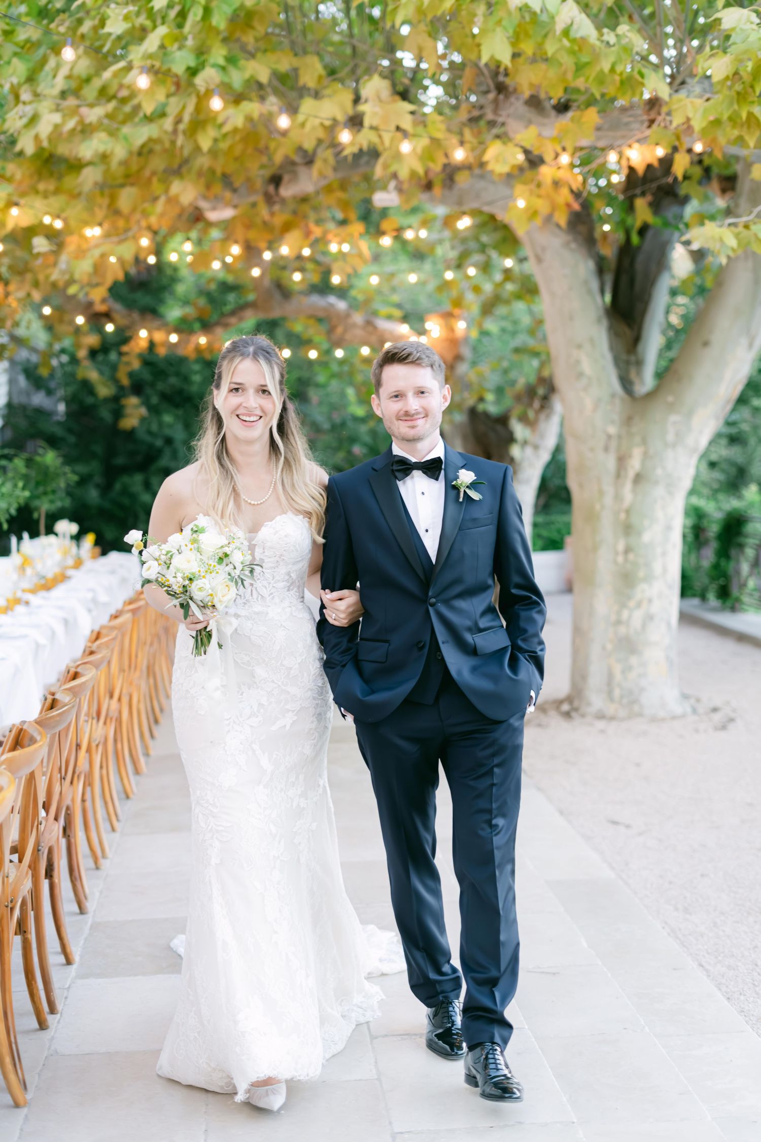 A couple walks together under autumn trees along a pathway lined with wooden chairs.