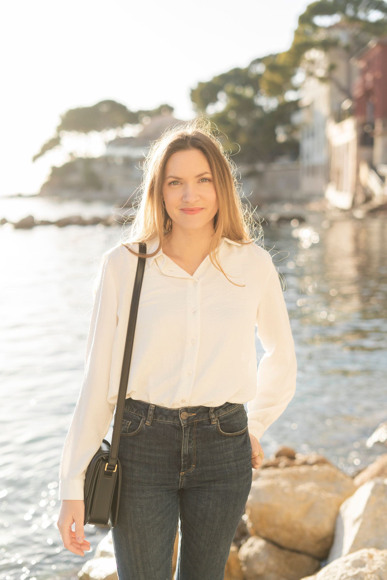 A person in a white blouse and jeans stands by a rocky coastline at sunset with sunlit water in the background.