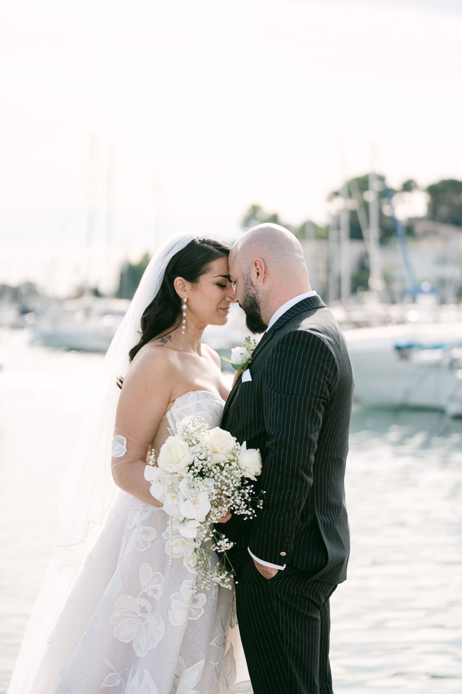 A bride and groom share an intimate moment by a marina with boats and white flowers in a dreamy seaside setting.