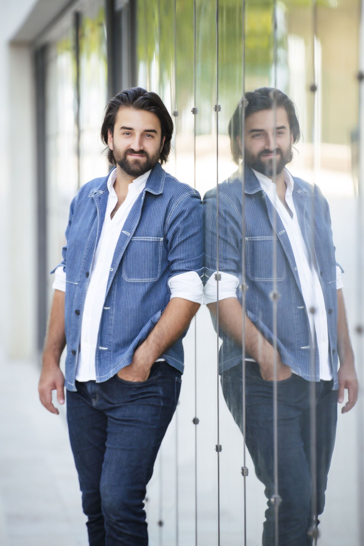 A man in a denim shirt leans against a reflective surface creating a mirrored image in casual professional attire.