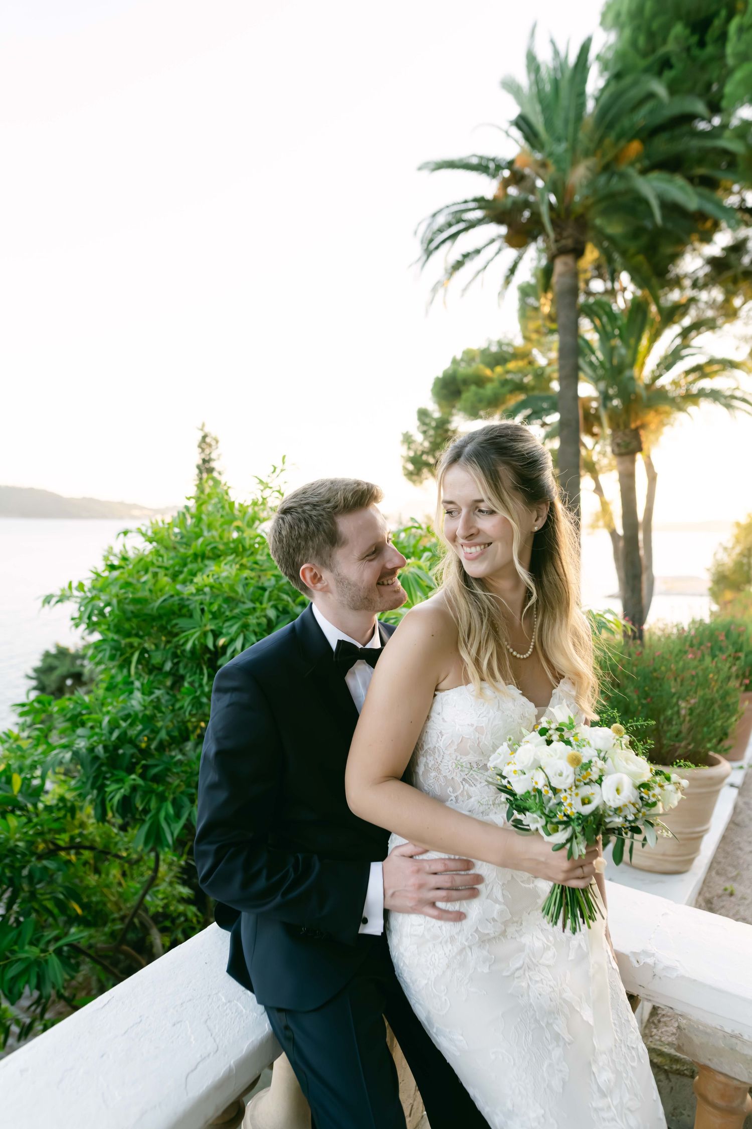 A romantic wedding couple embraces on a white terrace with palm trees and ocean views at sunset.