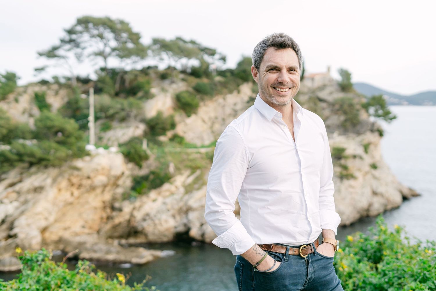 A person in a white shirt poses against a scenic coastal backdrop with rocky cliffs and ocean views.