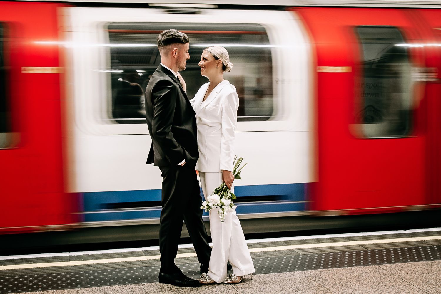 Couple in formal attire posing on London Underground platform with red train.