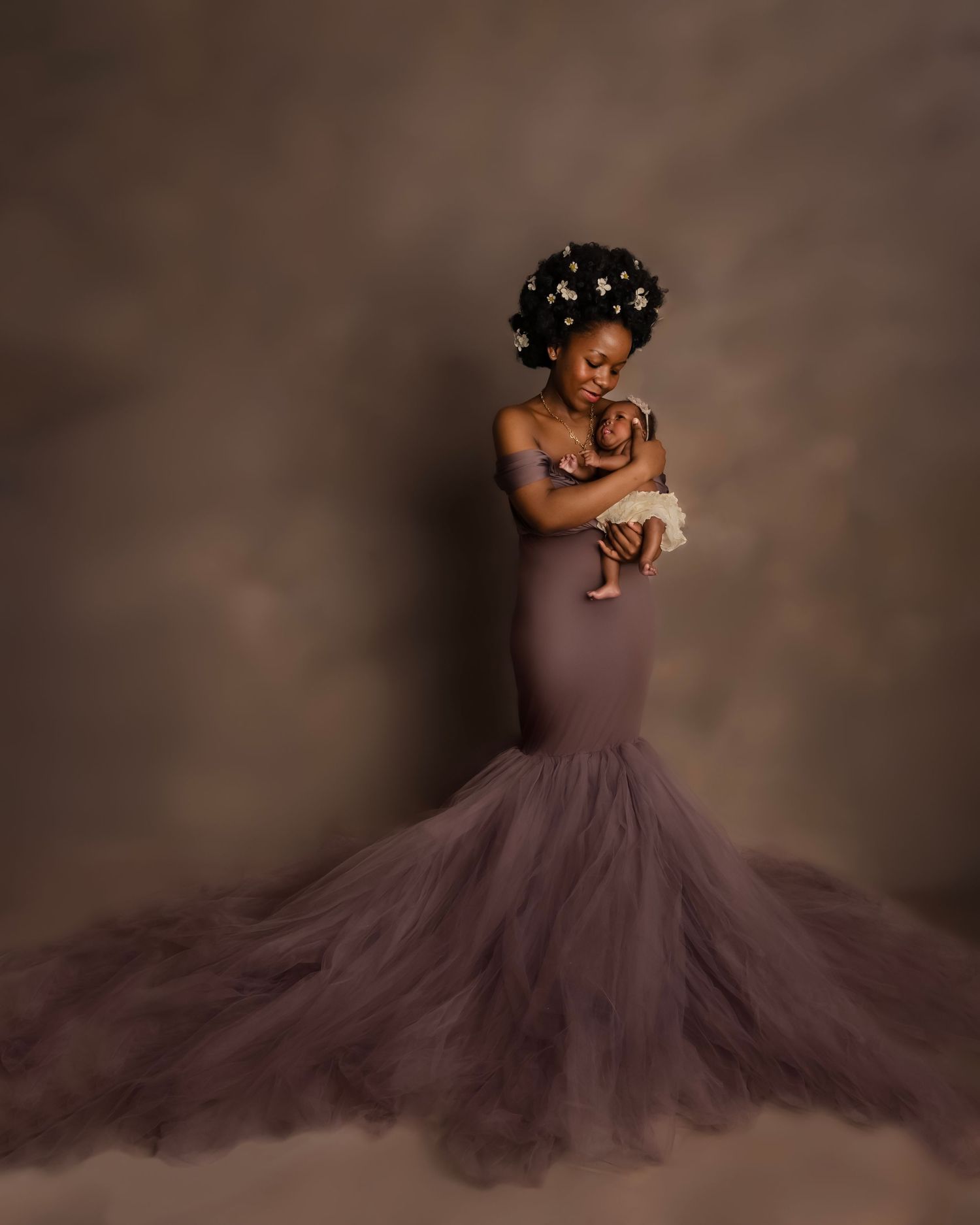 A woman in a purple tulle mermaid gown holds her newborn baby, wearing floral hair accessories, posed in a studio.