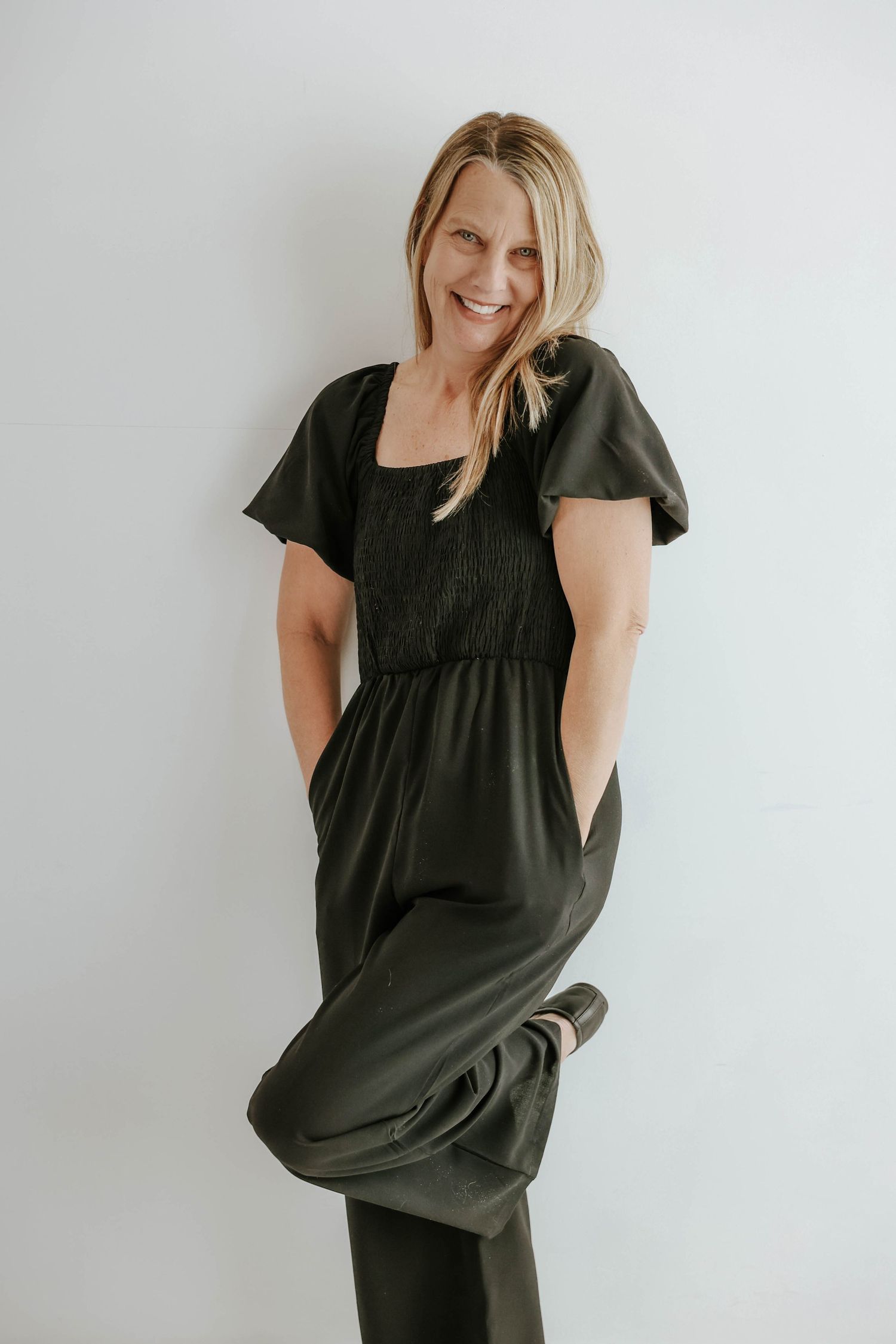 A person wearing a casual black maxi dress with short sleeves and pockets stands against a white background smiling.