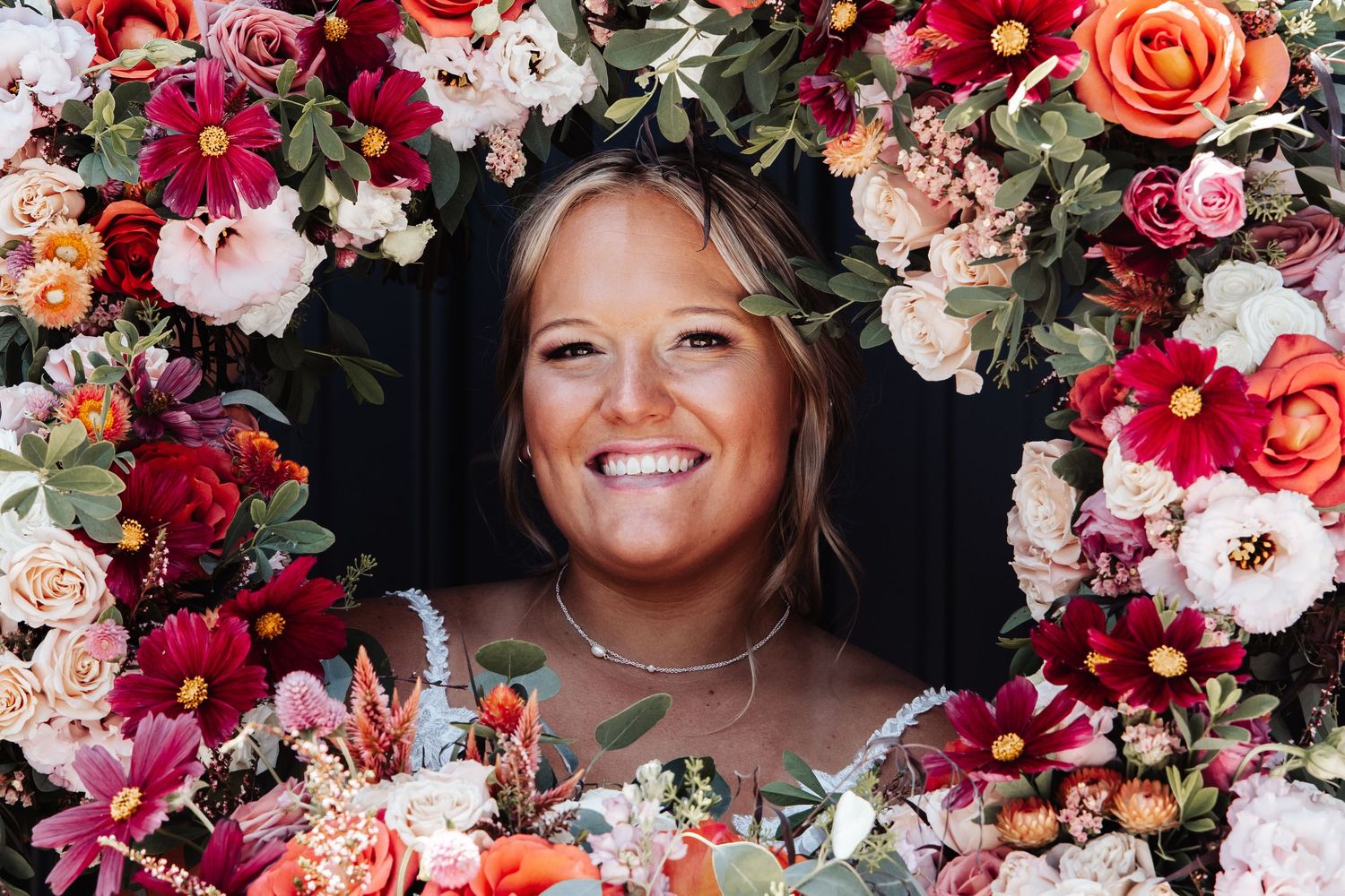 A portrait framed by a vibrant floral archway of pink, coral and red blooms with greenery against a dark background.