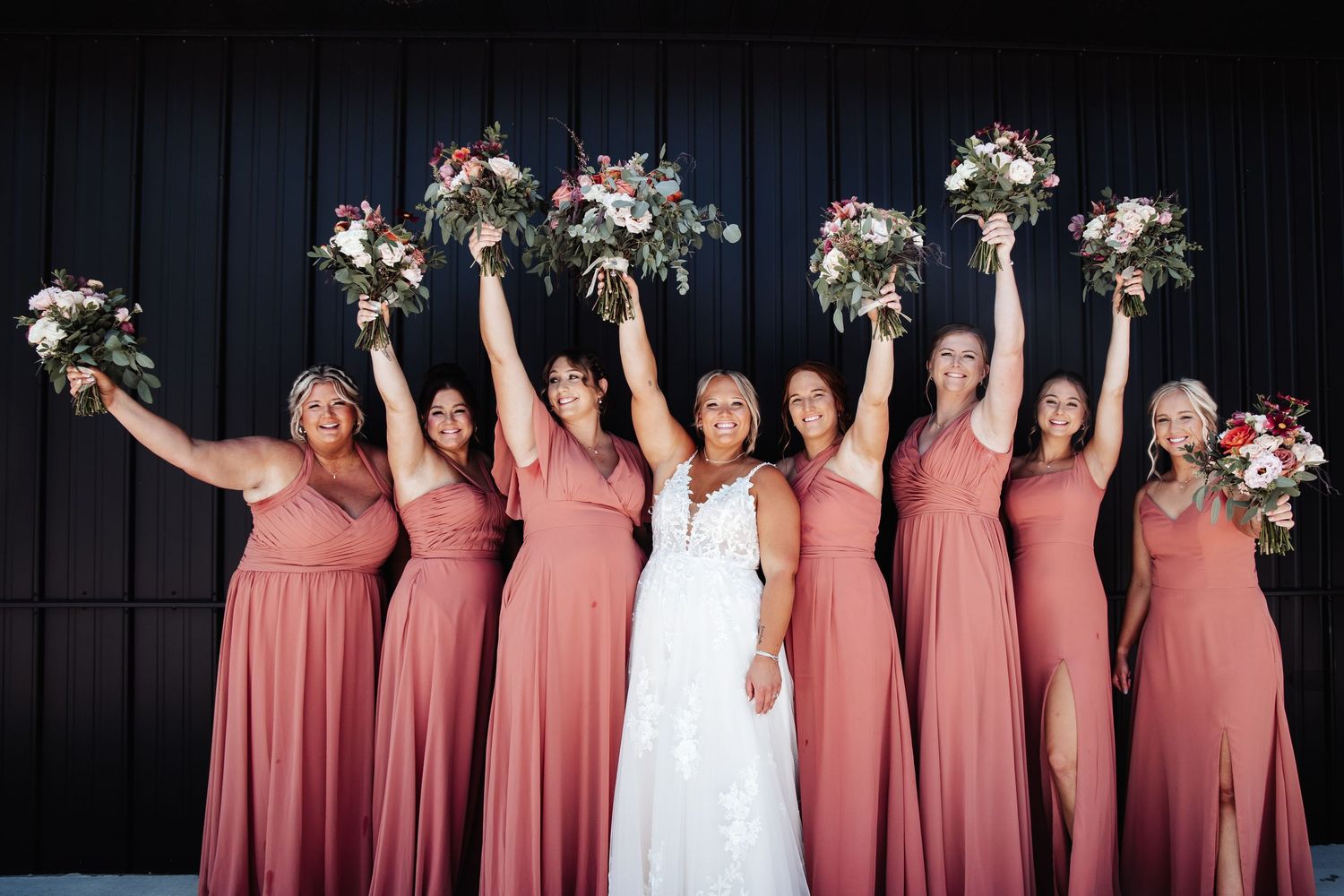 Wedding party in matching rose-colored dresses holding floral bouquets overhead against dark background.