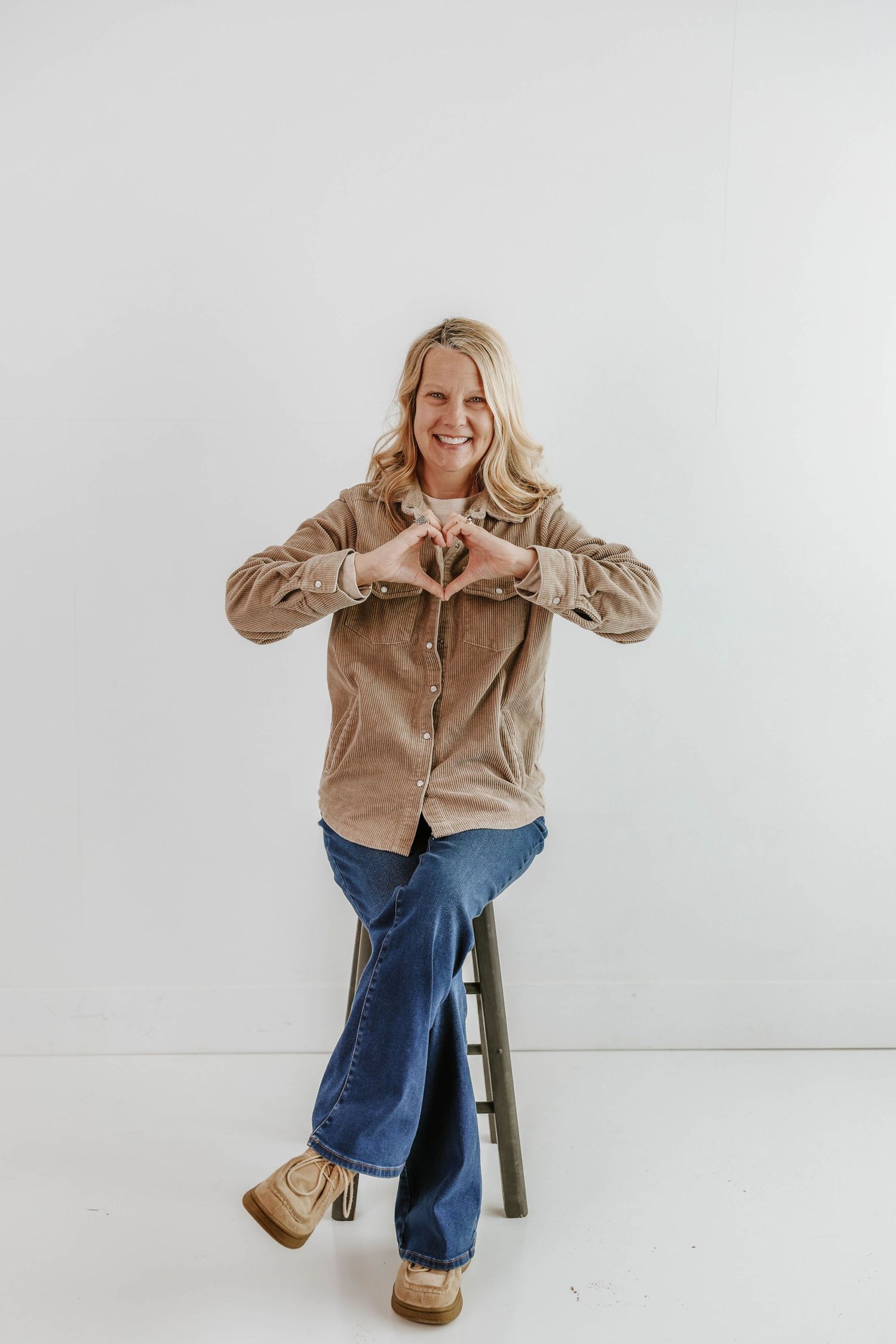 Casual portrait with heart hand gesture while sitting on wooden stool against white backdrop.