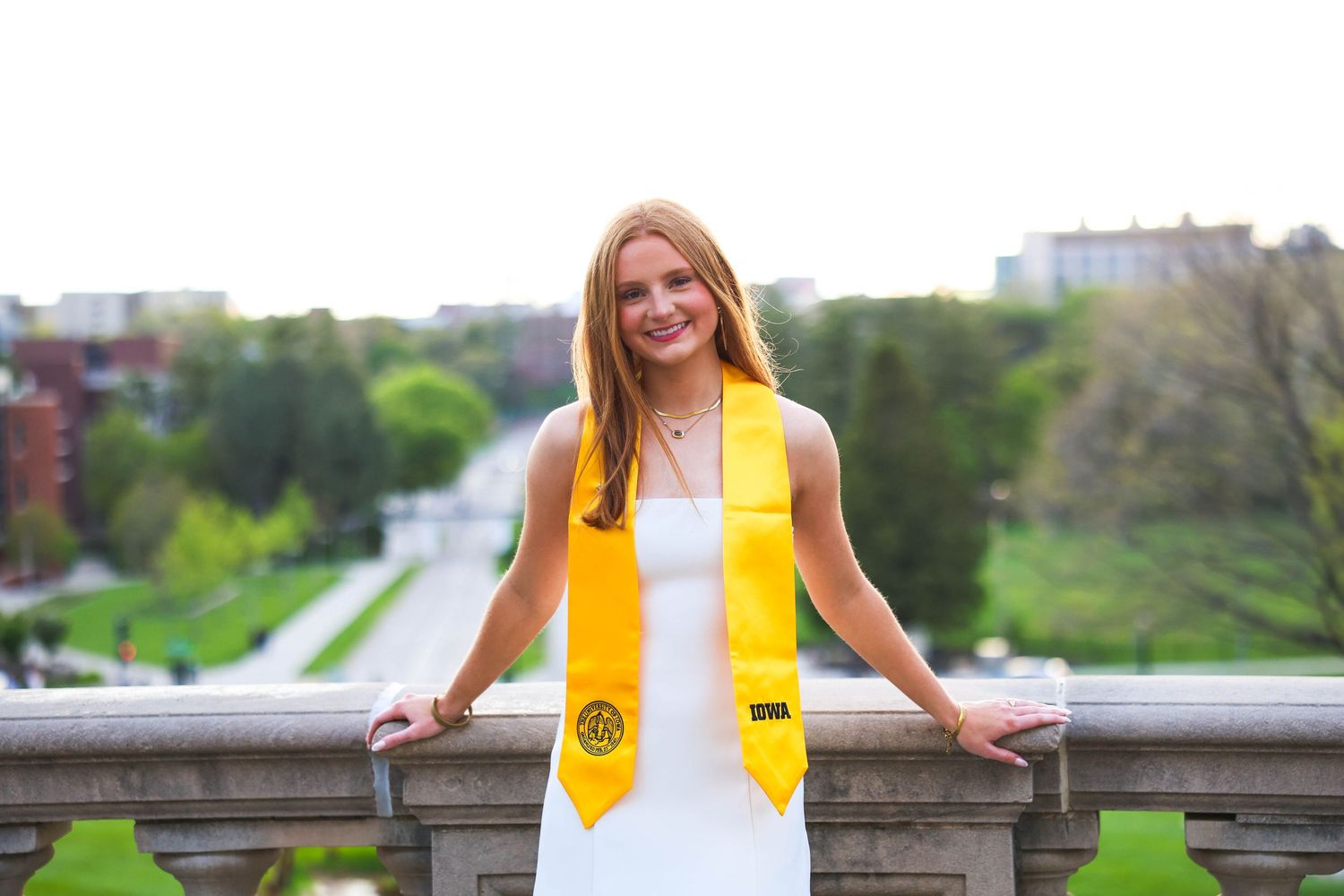 College graduate in white dress and yellow honors stole poses on stone balcony with trees in background.