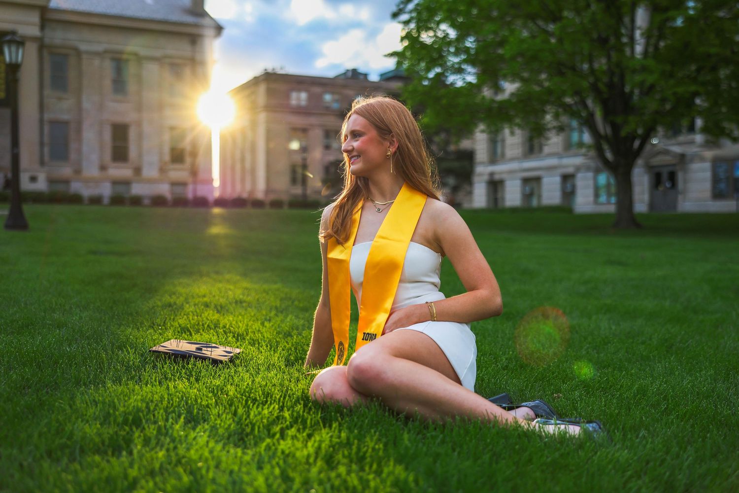 Graduate in white dress with yellow stole sits on sunny campus lawn at sunset with diploma beside her.